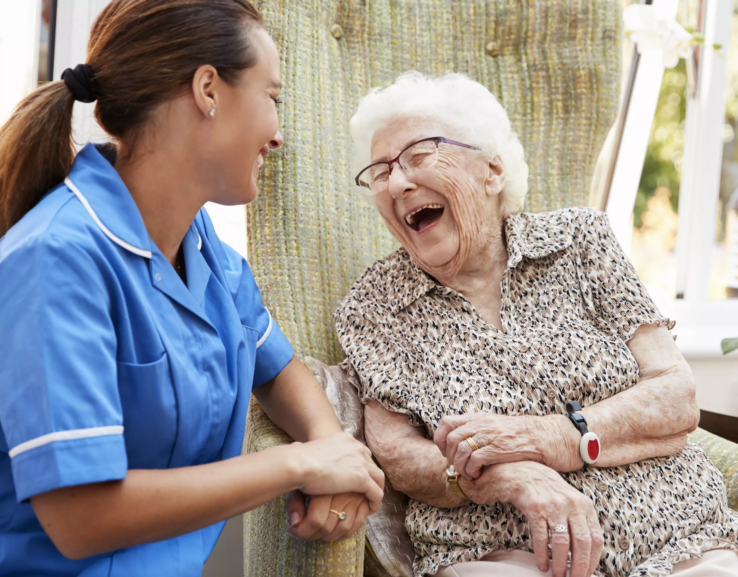 Ältere Dame sitzt im Rollsuthl und lacht mit einer Krankenpflegerin