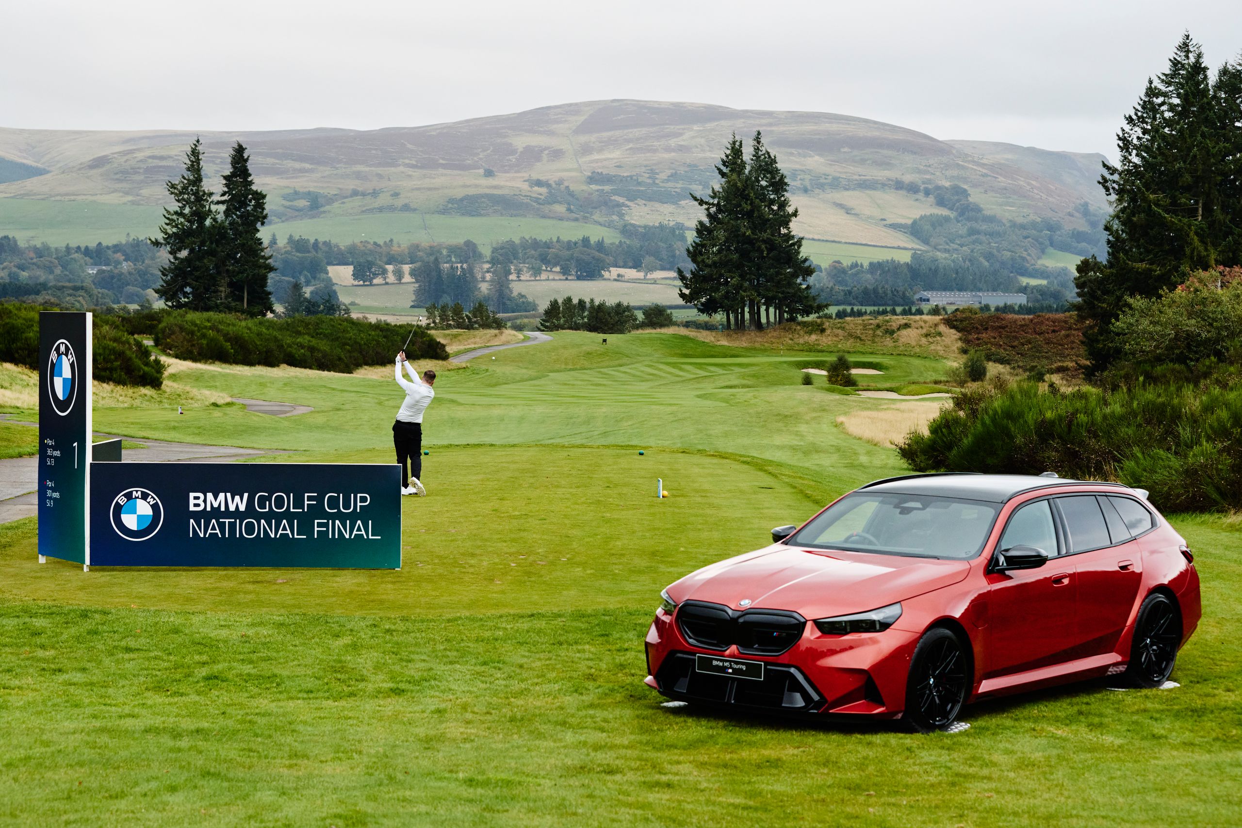 A golfer swings on a lush green course during the BMW Golf Cup National Final. A red BMW car is parked nearby, with scenic hills in the background.