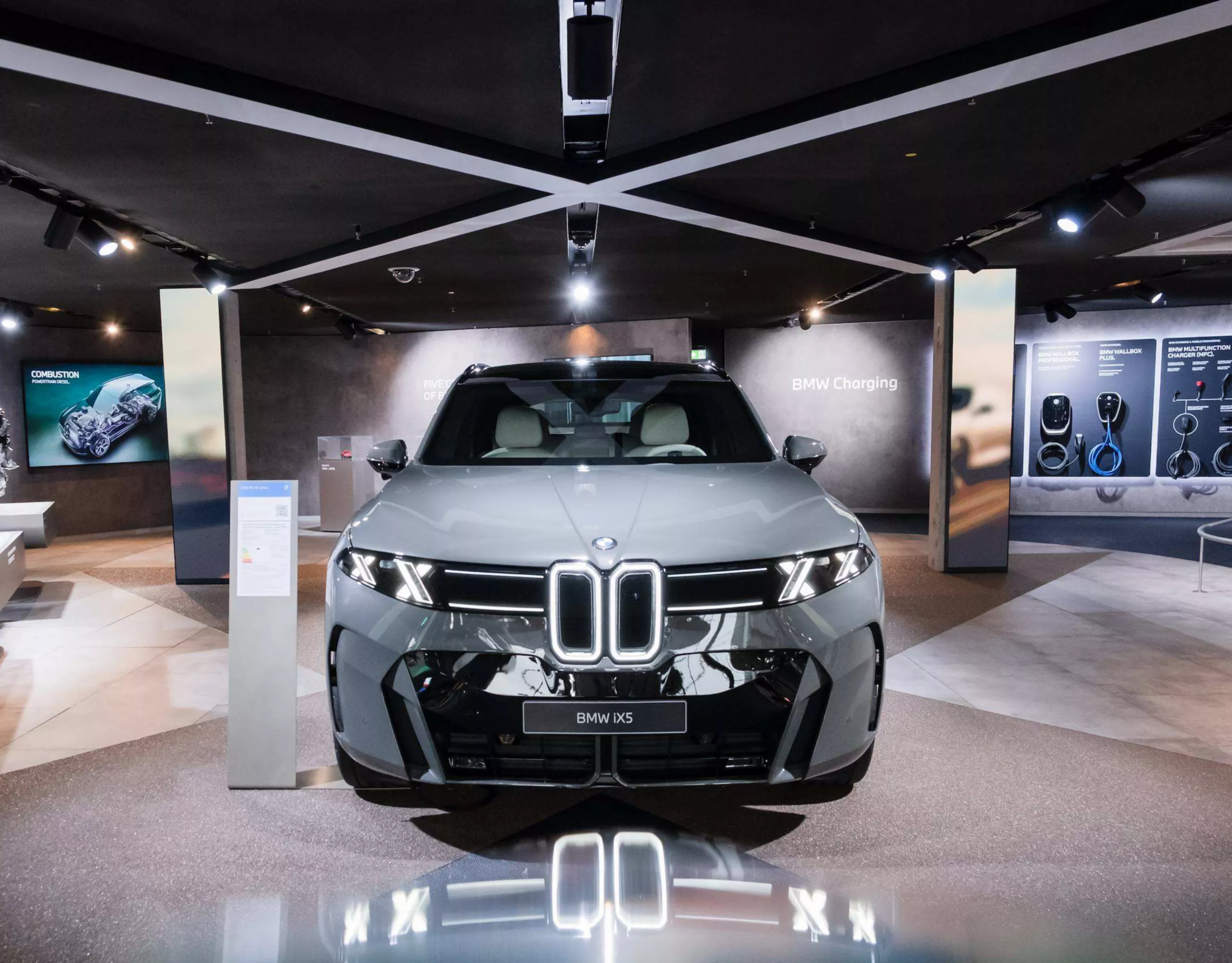 Frontansicht eines BMW iX5 in grey pine in einer modernen BMW-Ausstellung. Informationsflächen zu Ladetechnologien und Fahrzeugkomponenten umgeben das Fahrzeug.