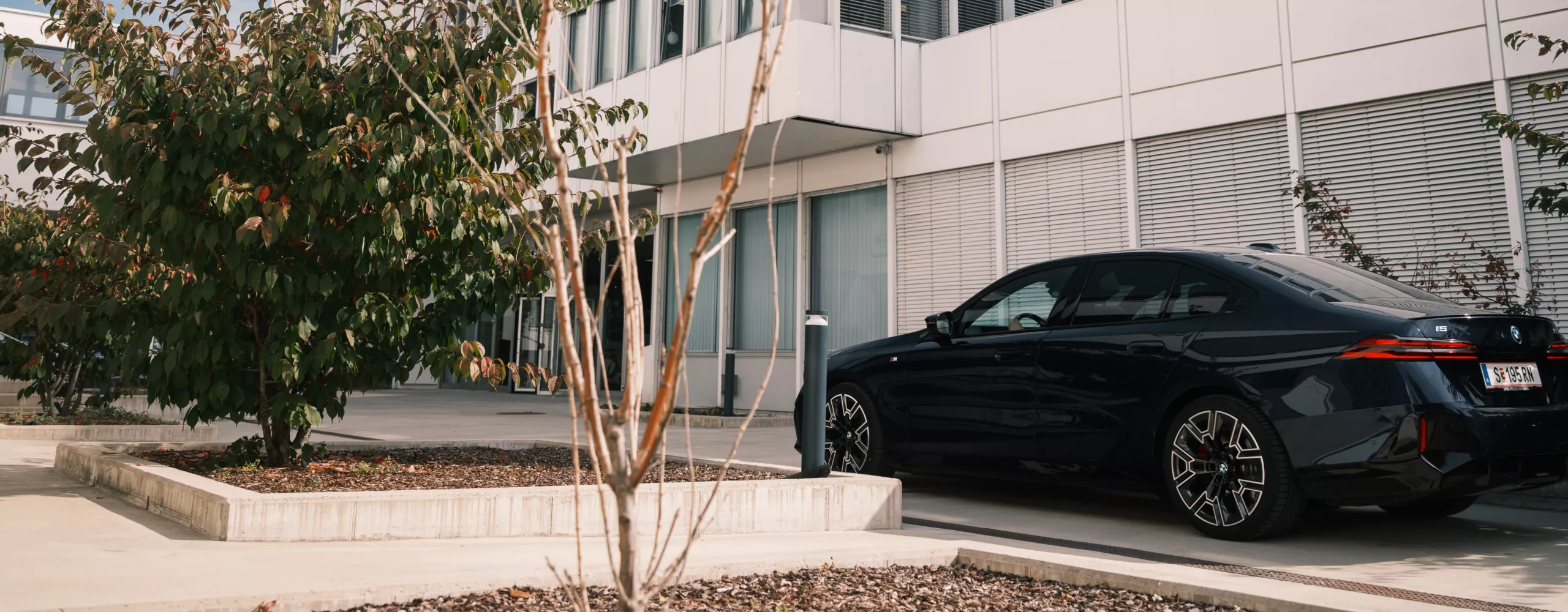 Ein dunkelbaluer BMW i5 steht vor dem BMW Group Standort Salzburg - Campus Süd. Das Gebäude besteht aus einer Fensterfront, an der teilweise die Jalousien heruntergelassen sind, und weißen Panelen. Im Vordergund stehen zwei Sträucher.