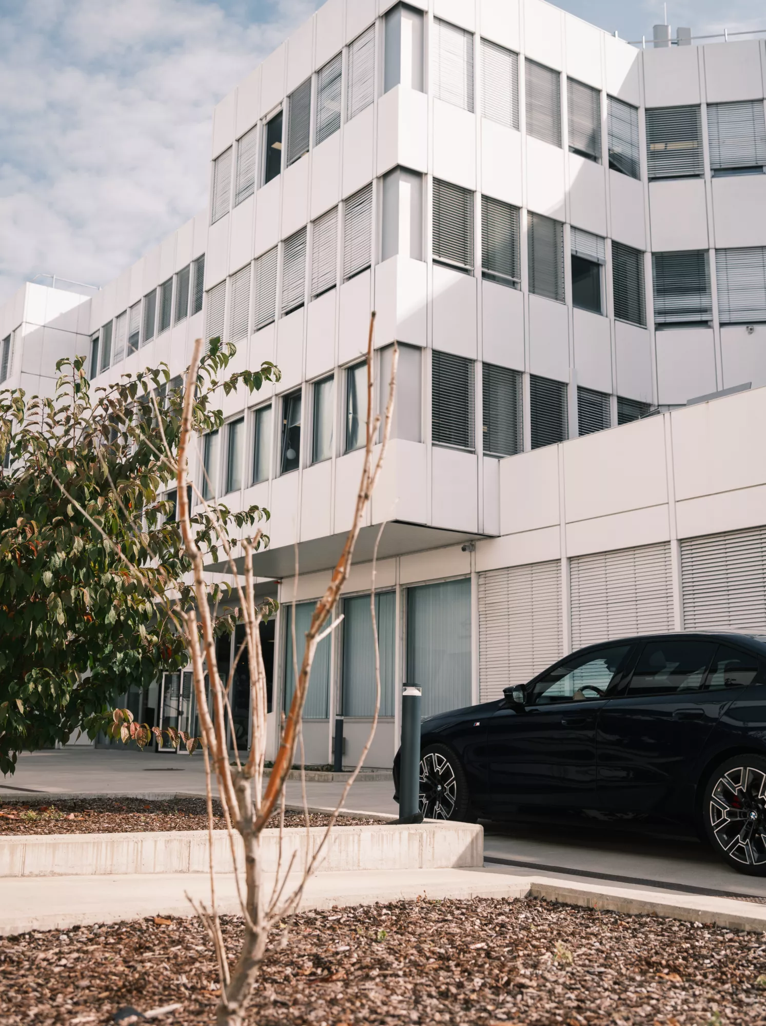 Ein dunkelbaluer BMW i5 steht vor dem BMW Group Standort Salzburg - Campus Süd. Das Gebäude besteht aus einer Fensterfront, an der teilweise die Jalousien heruntergelassen sind, und weißen Panelen. Im Vordergund stehen zwei Sträucher.