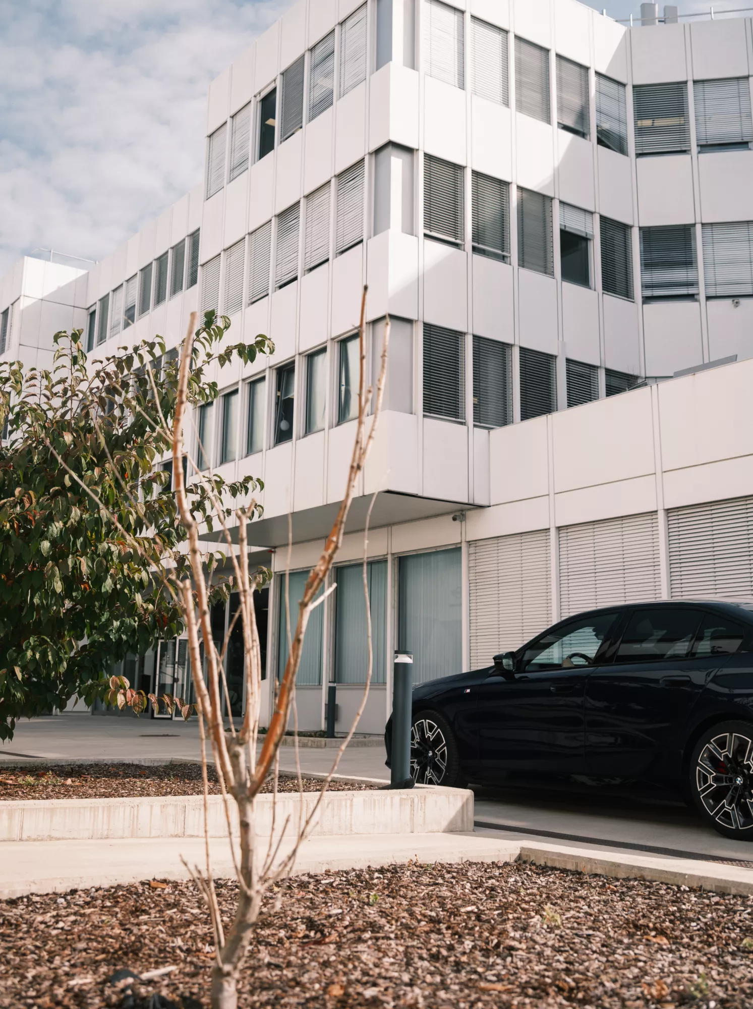 Ein dunkelbaluer BMW i5 steht vor dem BMW Group Standort Salzburg - Campus Süd. Das Gebäude besteht aus einer Fensterfront, an der teilweise die Jalousien heruntergelassen sind, und weißen Panelen. Im Vordergund stehen zwei Sträucher.