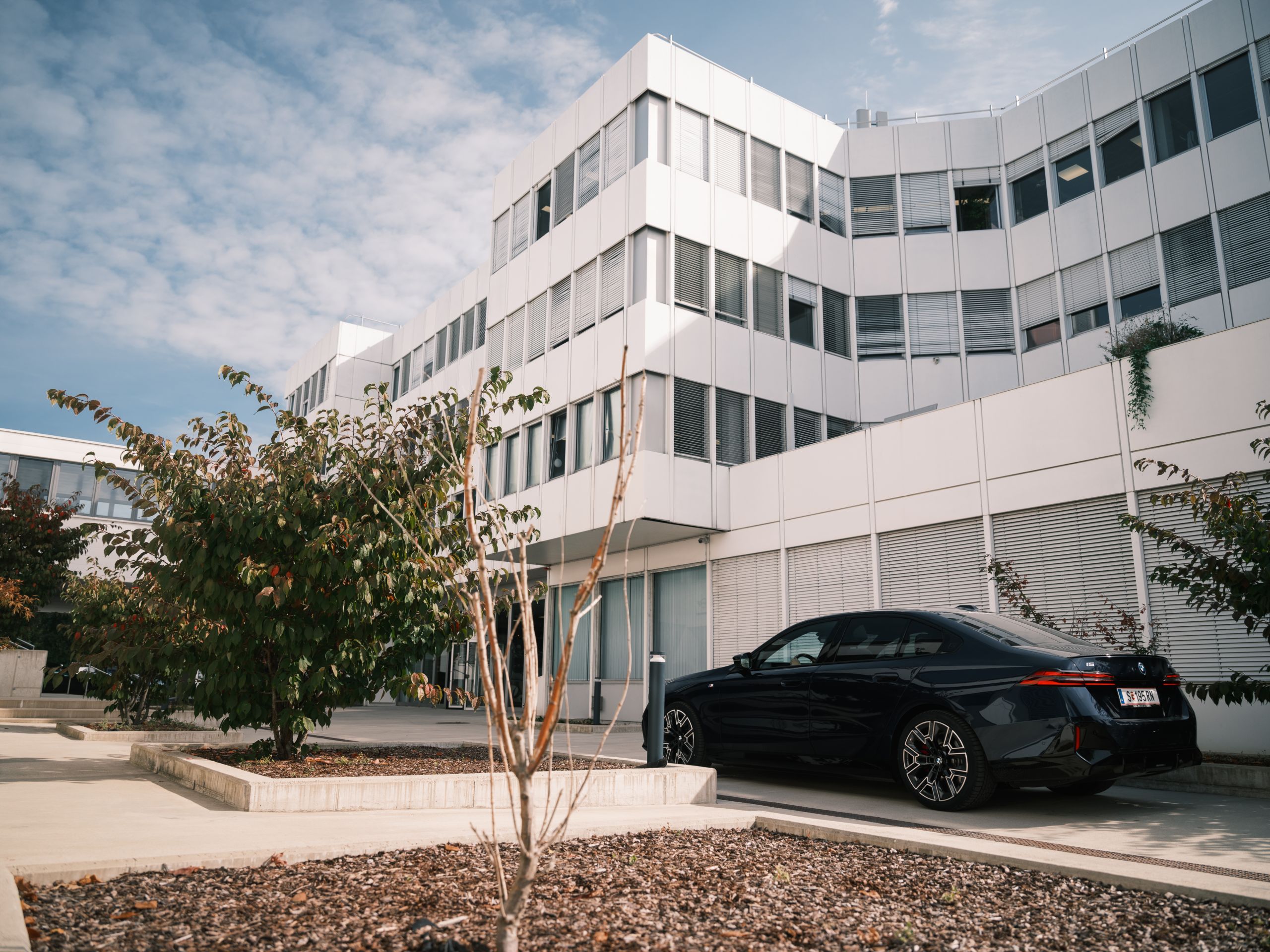 Ein dunkelbaluer BMW i5 steht vor dem BMW Group Standort Salzburg - Campus Süd. Das Gebäude besteht aus einer Fensterfront, an der teilweise die Jalousien heruntergelassen sind, und weißen Panelen. Im Vordergund stehen zwei Sträucher.