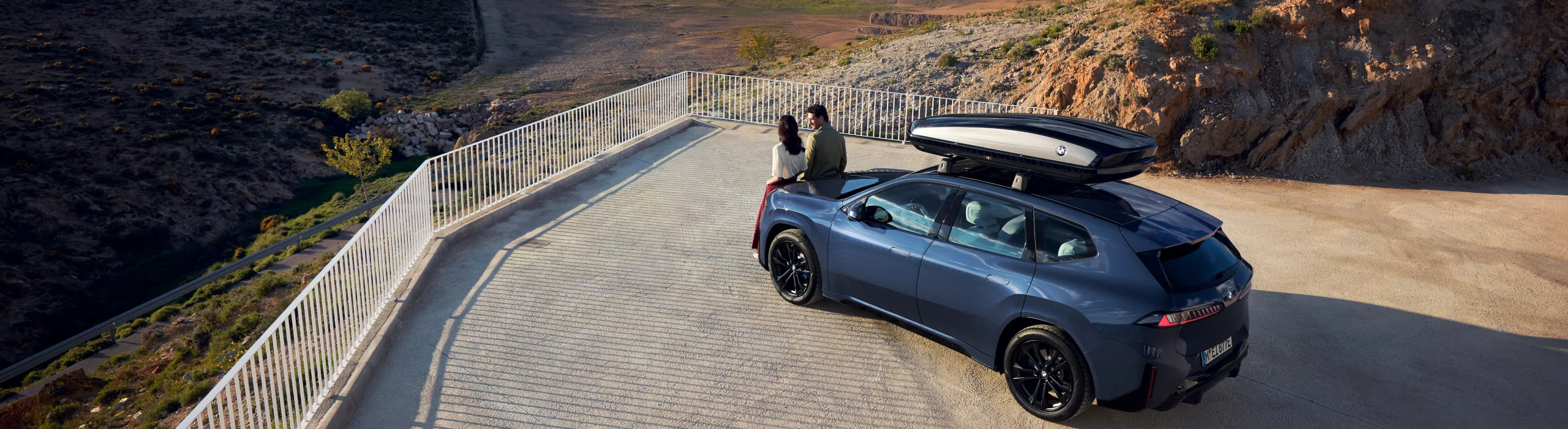 Zwei Personen stehen vor einem BMW iX3 mit Dachbox und blicken in die Ferne