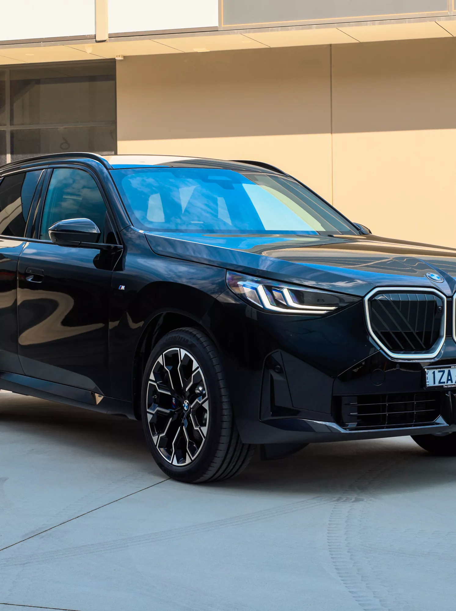A gloss black BMW X3 SUV parked on a concrete surface in front of a warm sandstone-coloured building, shot from a front three-quarter angle showcasing the illuminated kidney grille and M Sport styling details