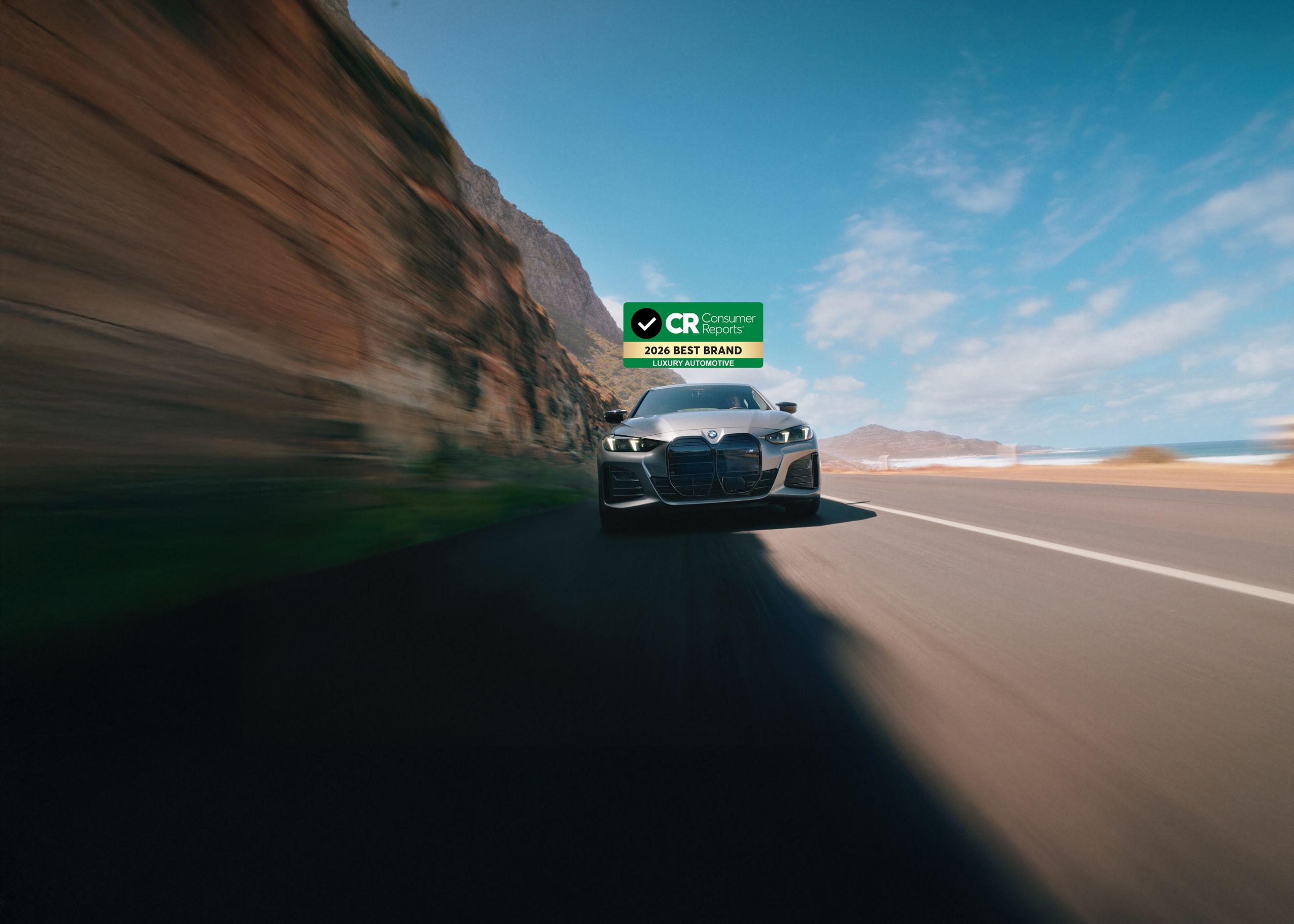 A front shot of a BMW i4 M50 as it drives along a canyon highway. The Consumer Reports 2026 Best Luxury automotive brand logo sits at the top of the image.