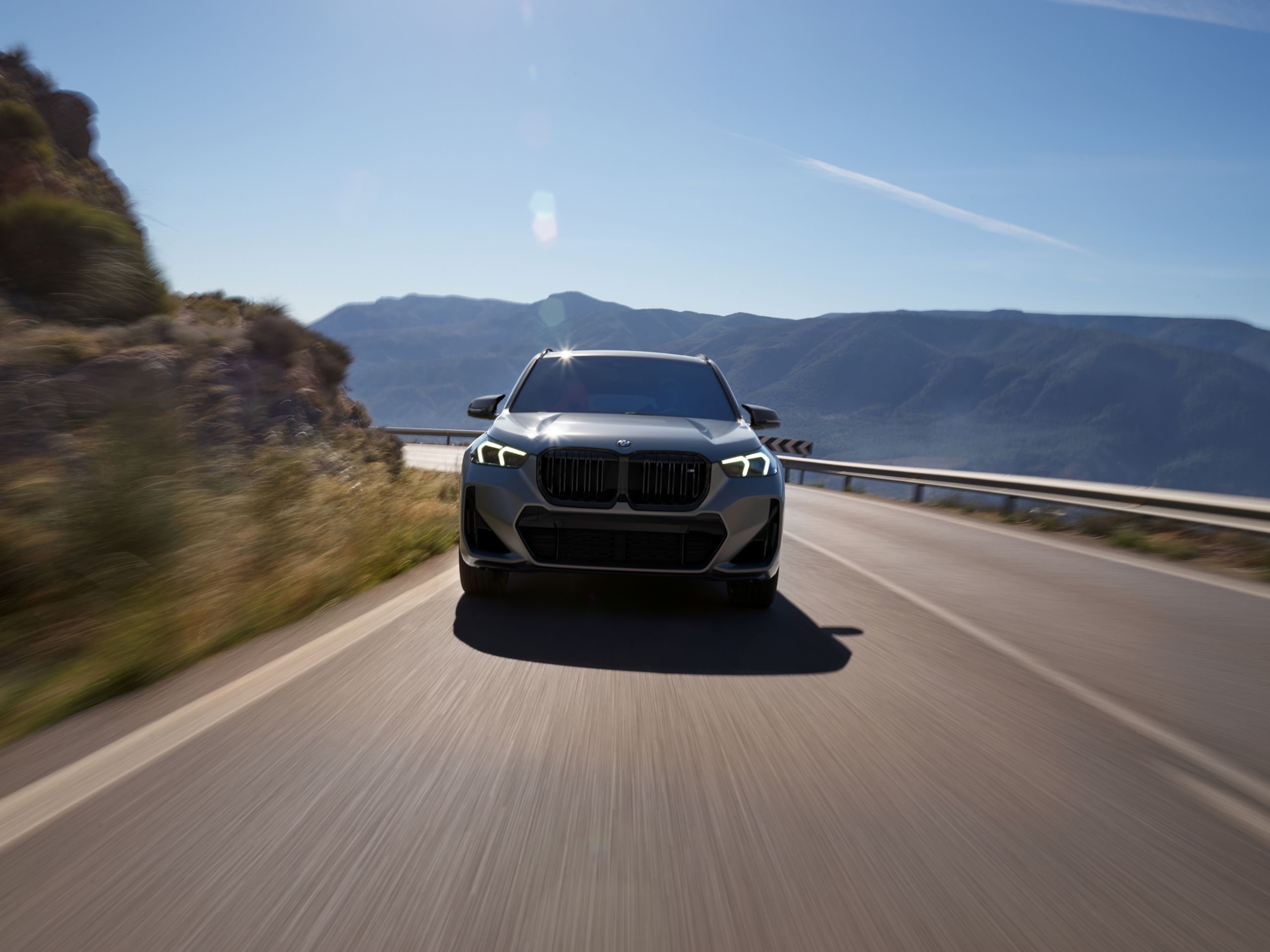 Front view of a matte gray 2026 BMW X1 M35i driving on a curvy road on a sunny day with mountains in the background.