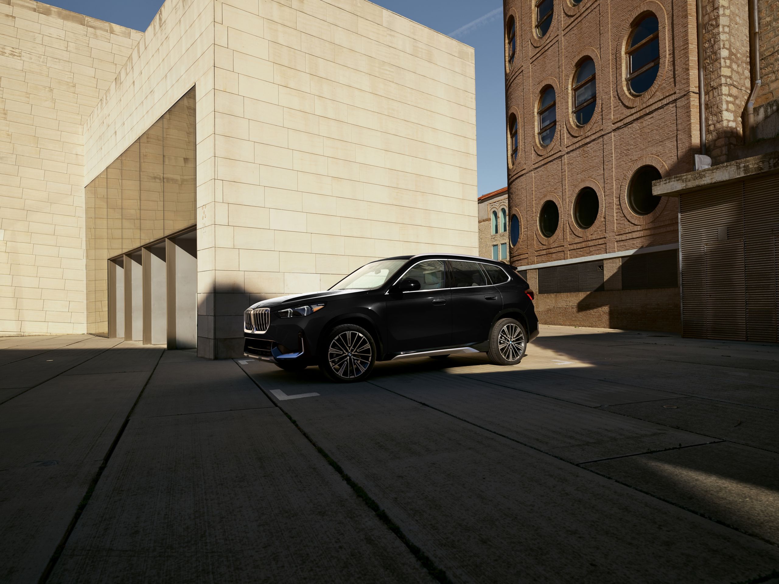 An BMW X1 xDrive28i SUV parked in a sunlit city scene in front of a beige brick wall. 