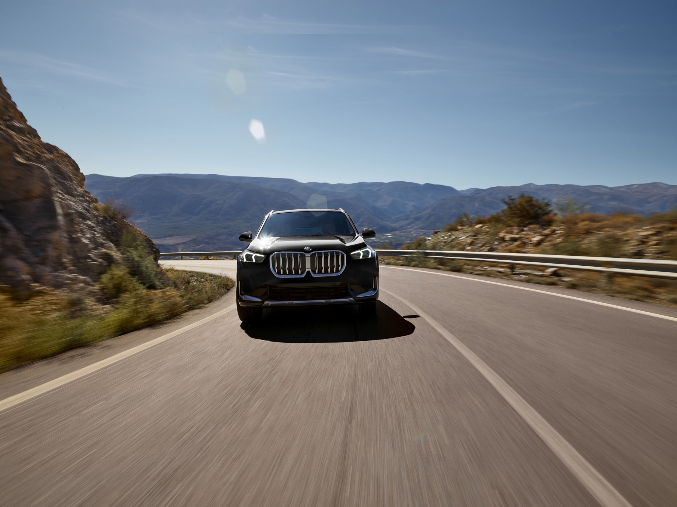 Front view of an 2026 BMW X1 driving on a curvy road on a sunny day with mountains in the background.