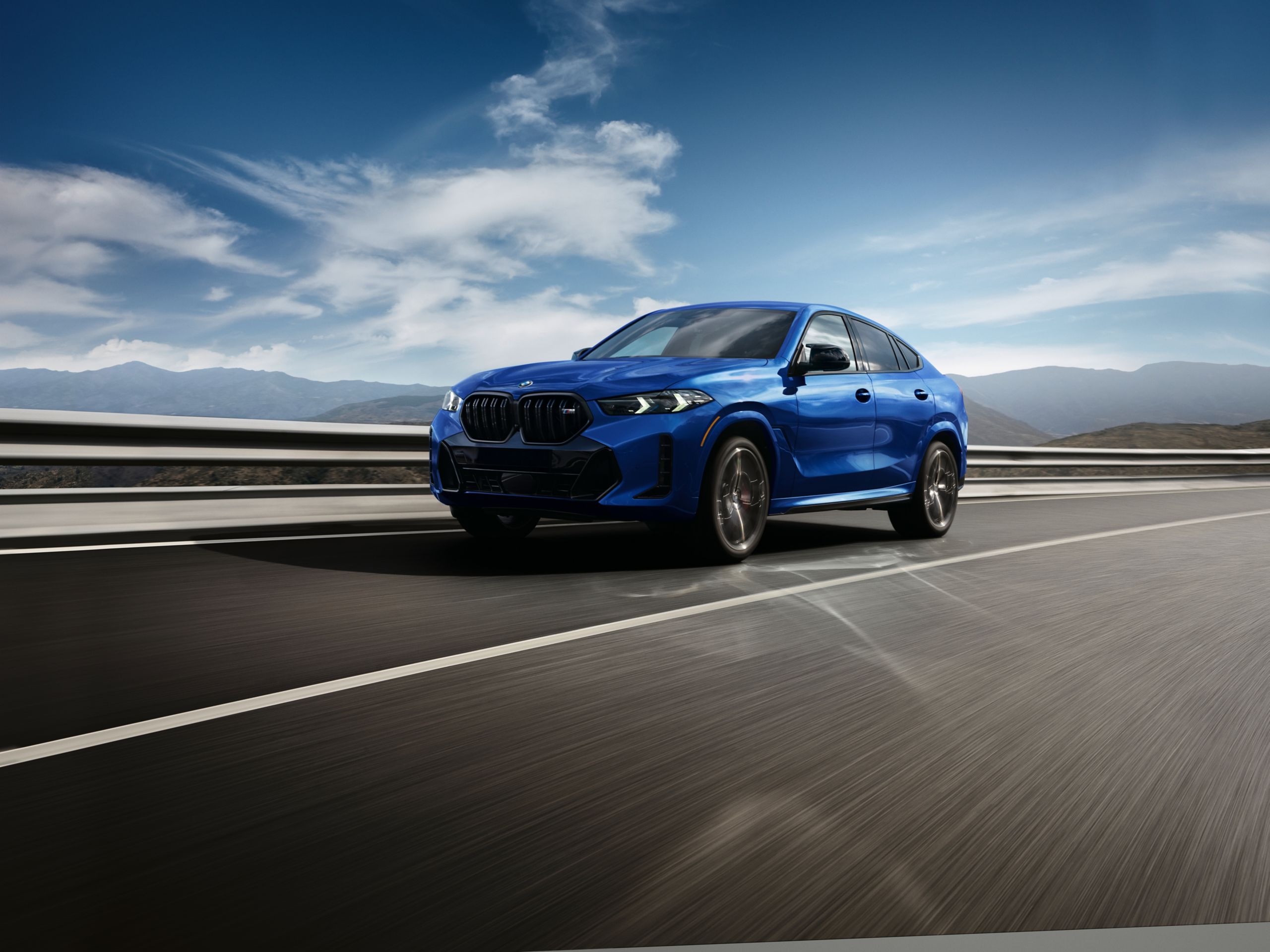 A BMW X6 M60i xDrive is driving at high speeds on a highway in front of a mountain panorama