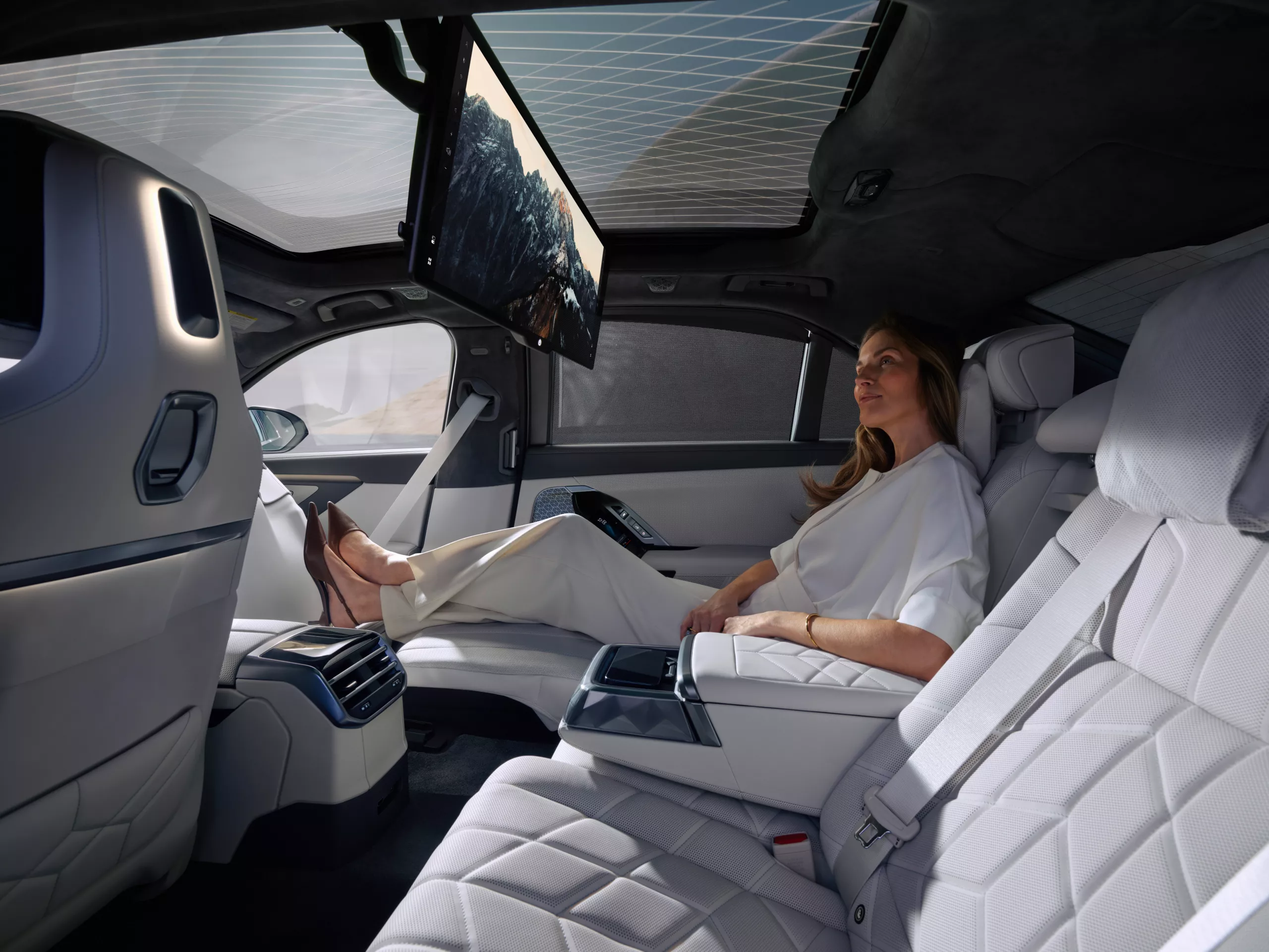 A passenger sits in the rear lounge seating of a BMW with her legs up, looking at a large passenger screen showing an image of a mountain range. 
