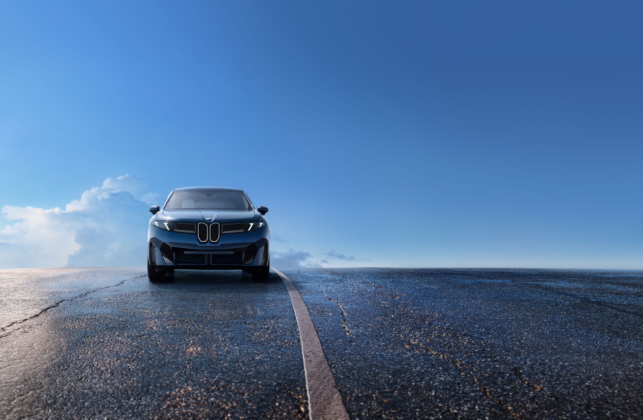 A BMW iX3 is driving on a wet, elevated road in front of a clear blue sky with few clouds 