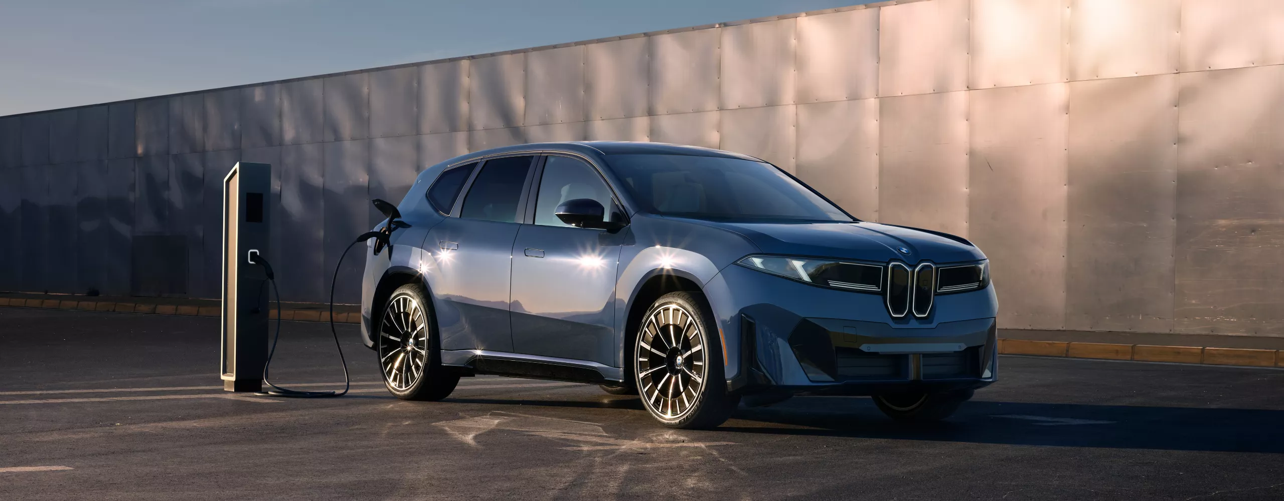 A BMW iX3 plugged into a charging station in front of a large, paneled metal wall.