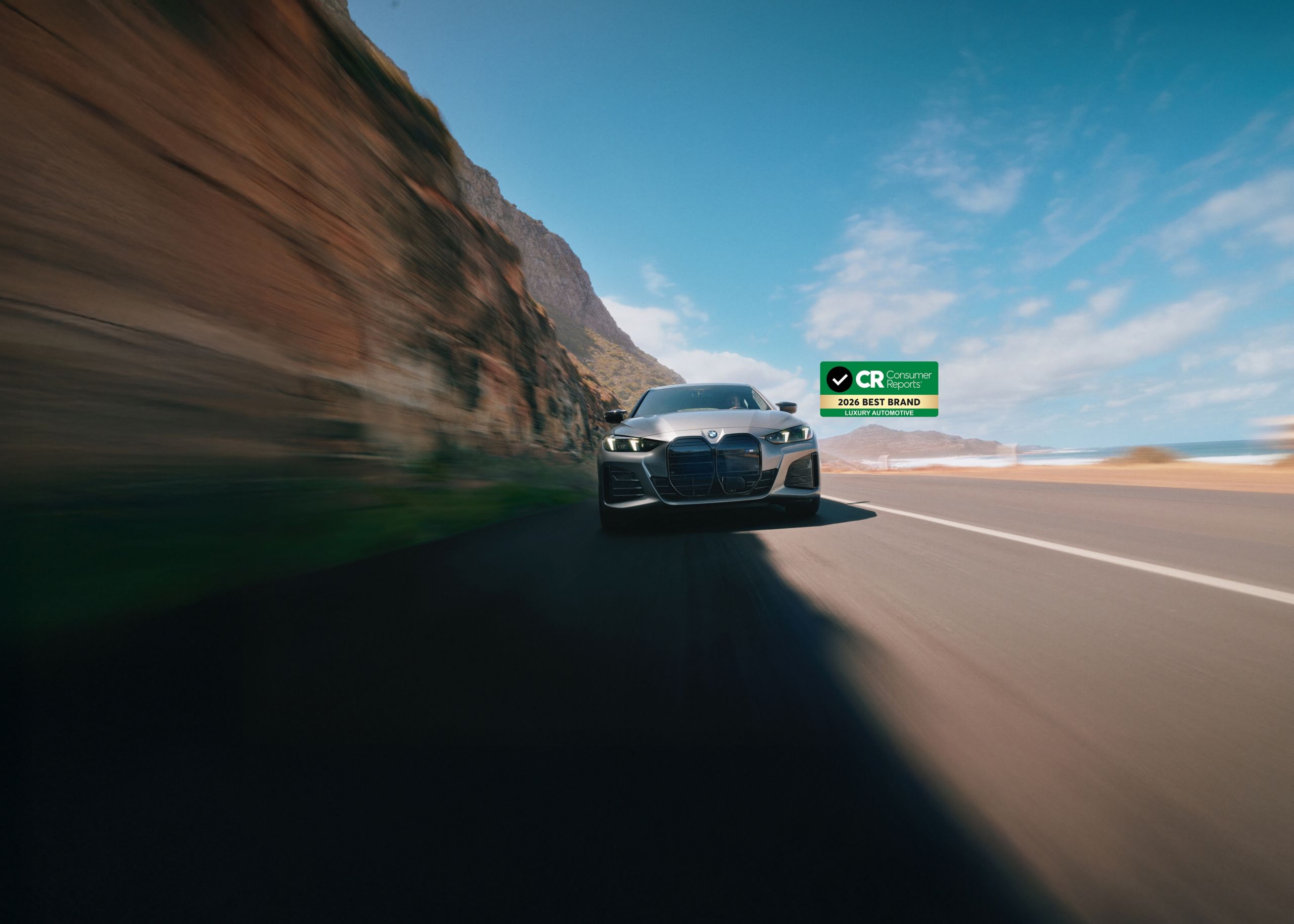 A front shot of a BMW i4 M50 as it drives along a canyon highway. The Consumer Reports 2026 Best Luxury automotive brand logo sits at the top of the image.