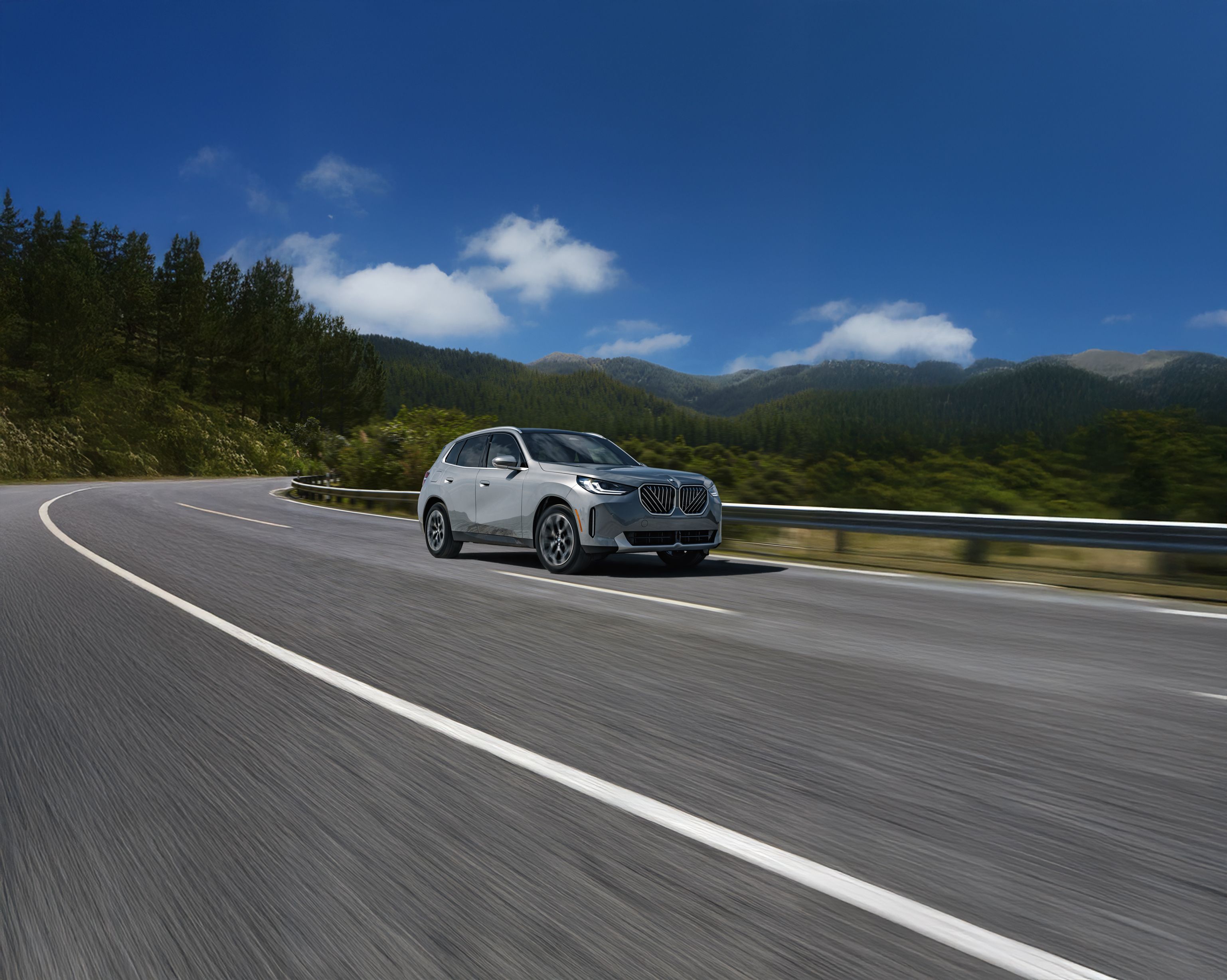 The BMW X3 driving along a winding road surrounded by a mountainous, wooded landscape under a partly cloudy sky.