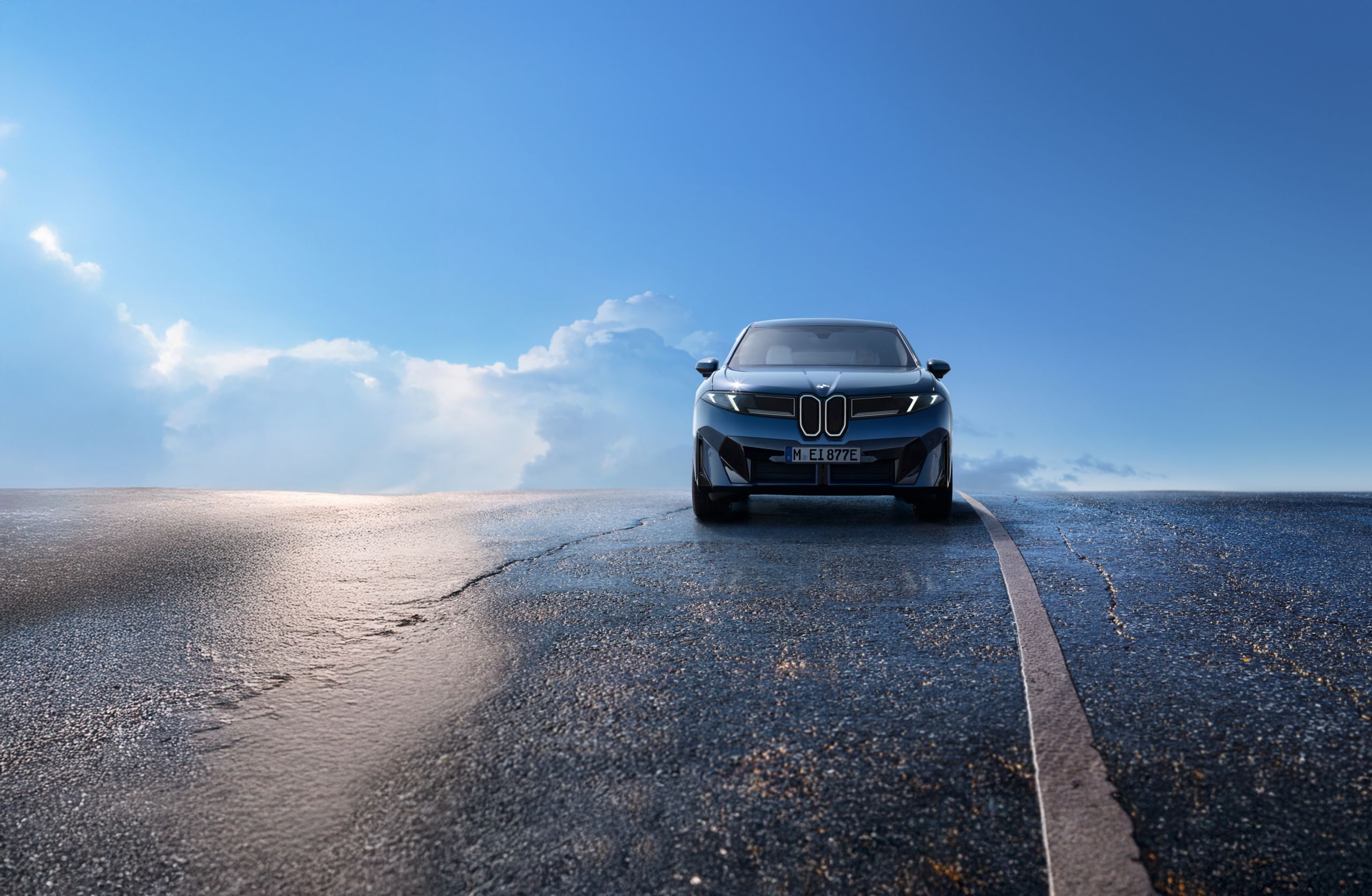 The 2027 BMW iX3 posed powerfully on an asphalt road, directly facing the viewer, with a blue sky in the background.
