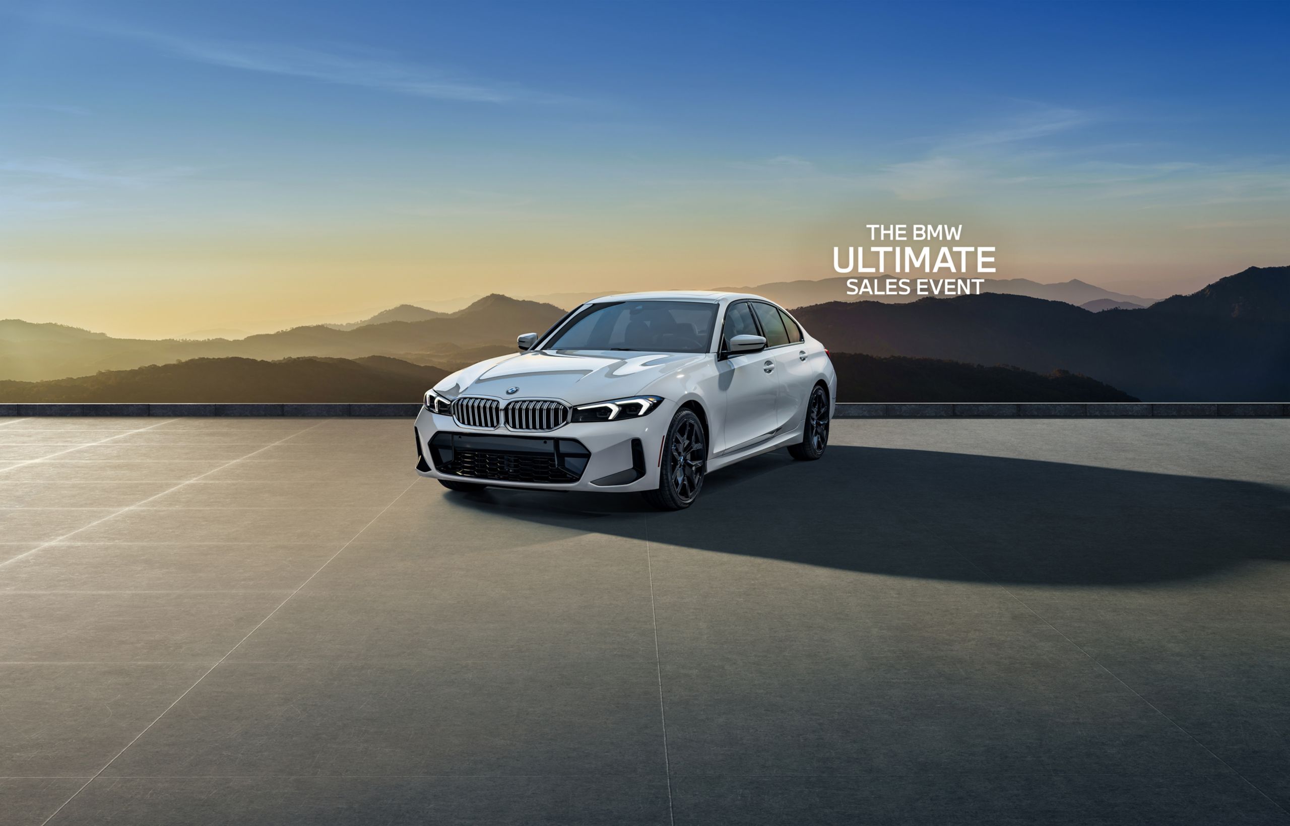 The BMW 3 Series parked on a paved area in front of a silhouetted mountain landscape at dawn or dusk.