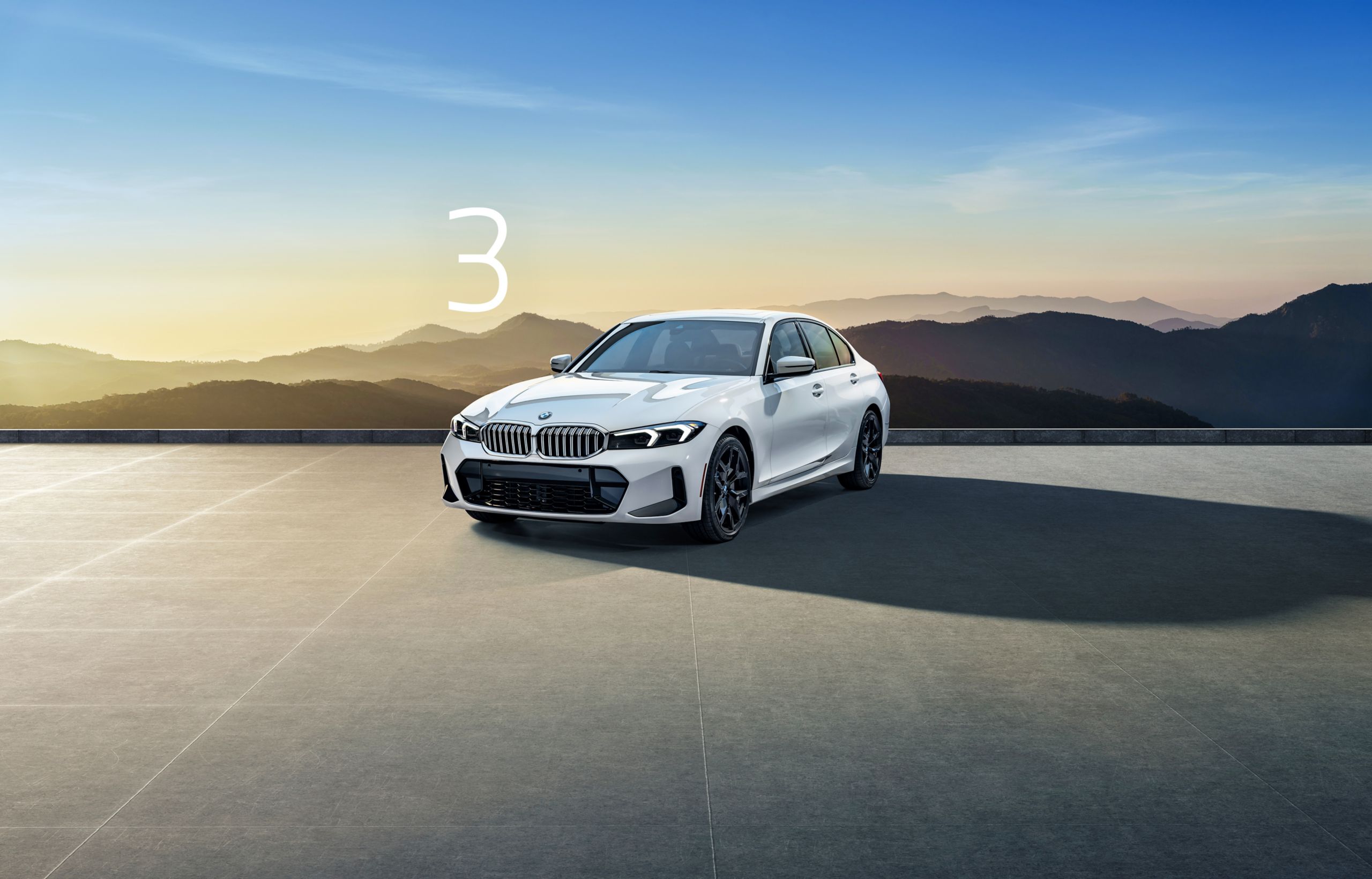 The BMW 3 Series parked on a paved area in front of a silhouetted mountain landscape at dawn or dusk.