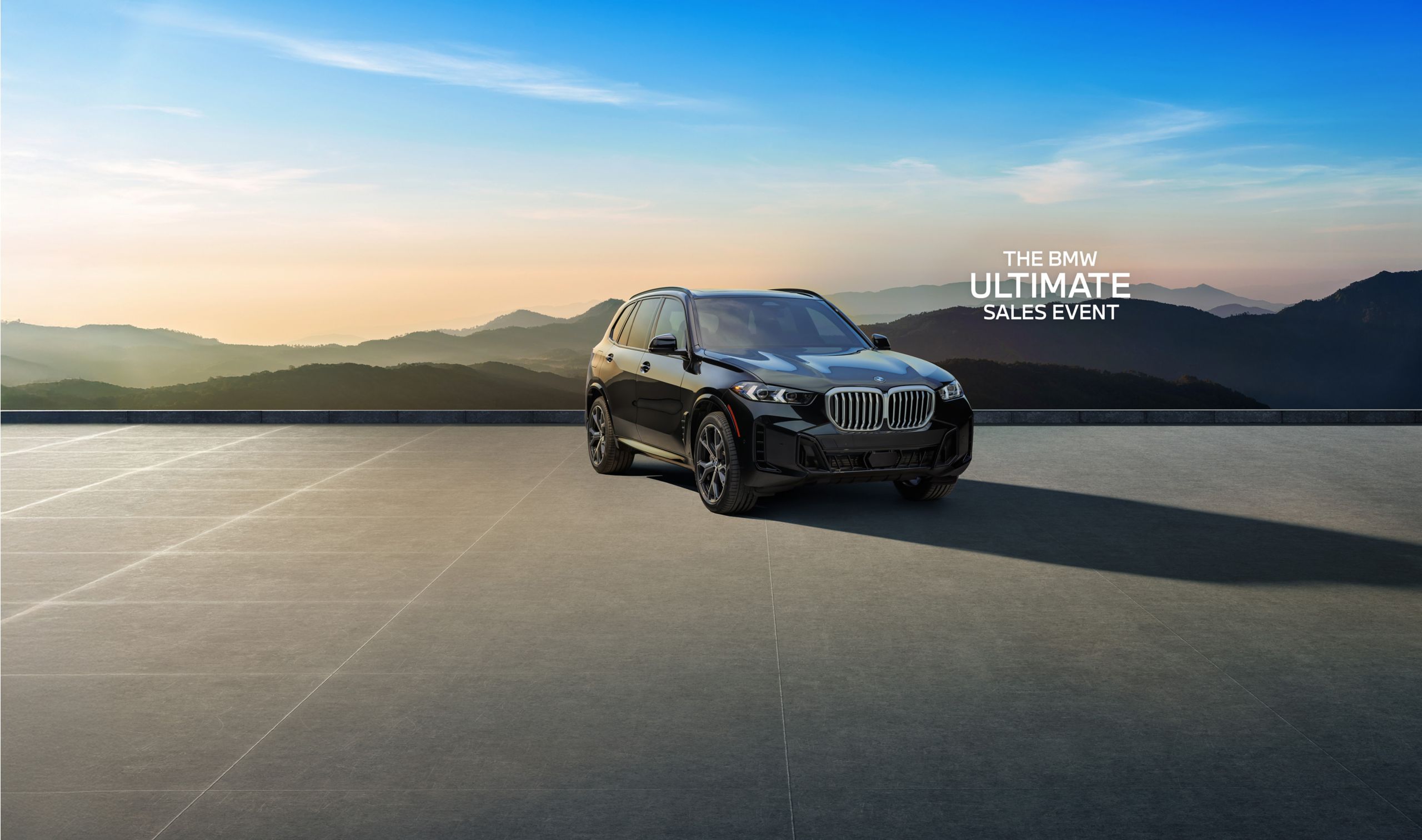 The BMW X5 parked on a paved area in front of a silhouetted mountain landscape at dawn or dusk.