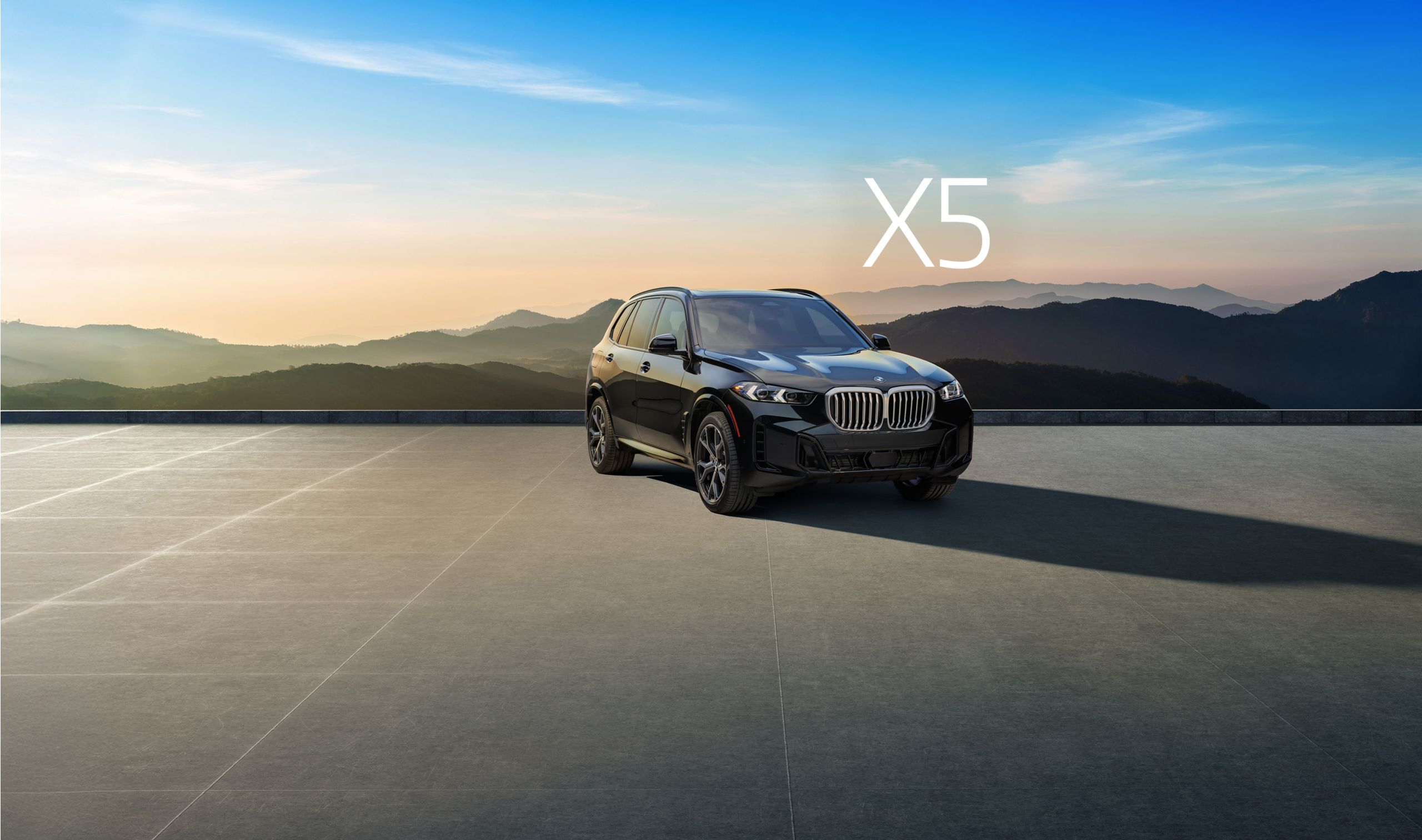 The BMW X5 parked on a paved area in front of a silhouetted mountain landscape at dawn or dusk.