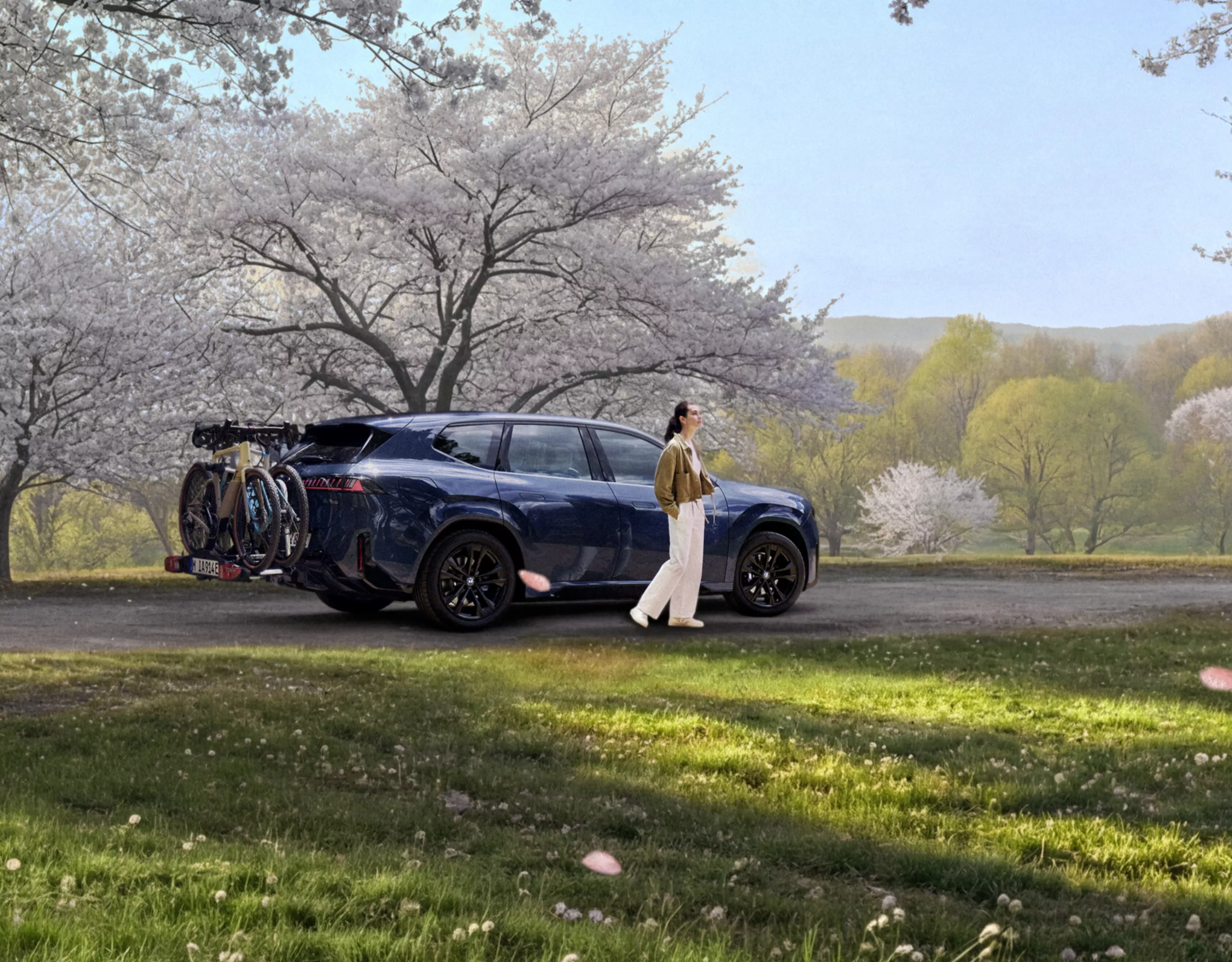 BMW iX3 (NA5) con bicicletas en el portabicicletas trasero, estacionado en un entorno natural con árboles en flor, mientras una persona camina a su lado