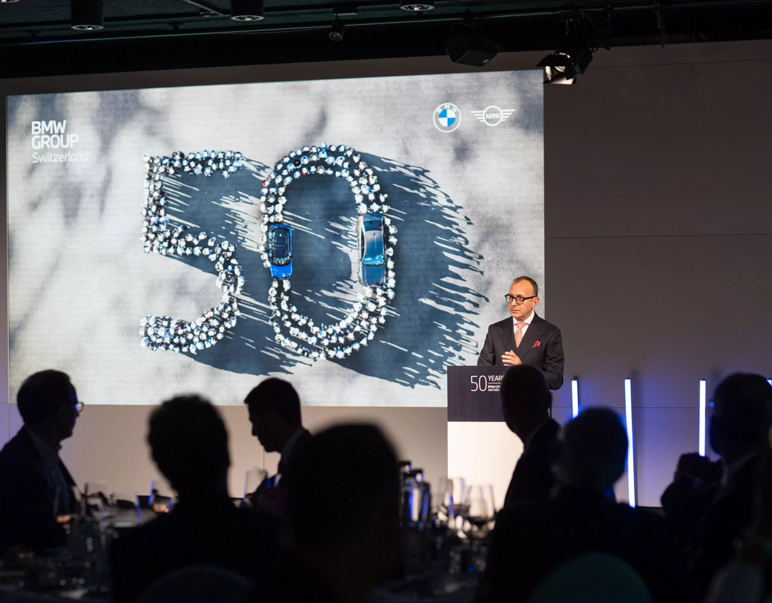 Un uomo su un palcoscenico davanti al podio dell’oratore con una presentazione sui 50 anni del BMW Group Switzerland sullo sfondo