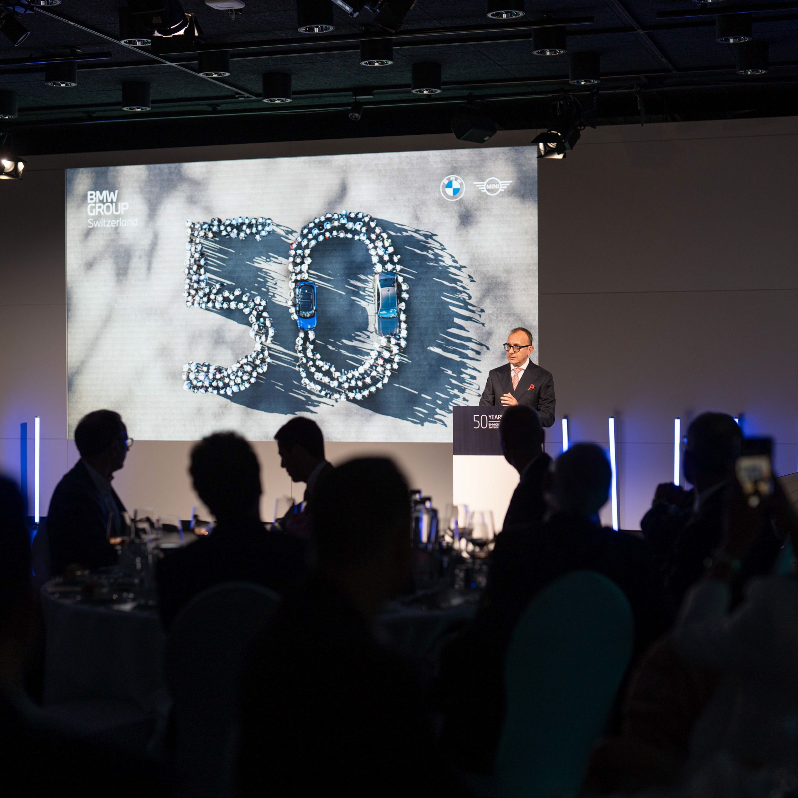 Un homme se tient sur une scène devant un pupitre avec une présentation sur les 50 ans du BMW Group Switzerland en arrière-plan