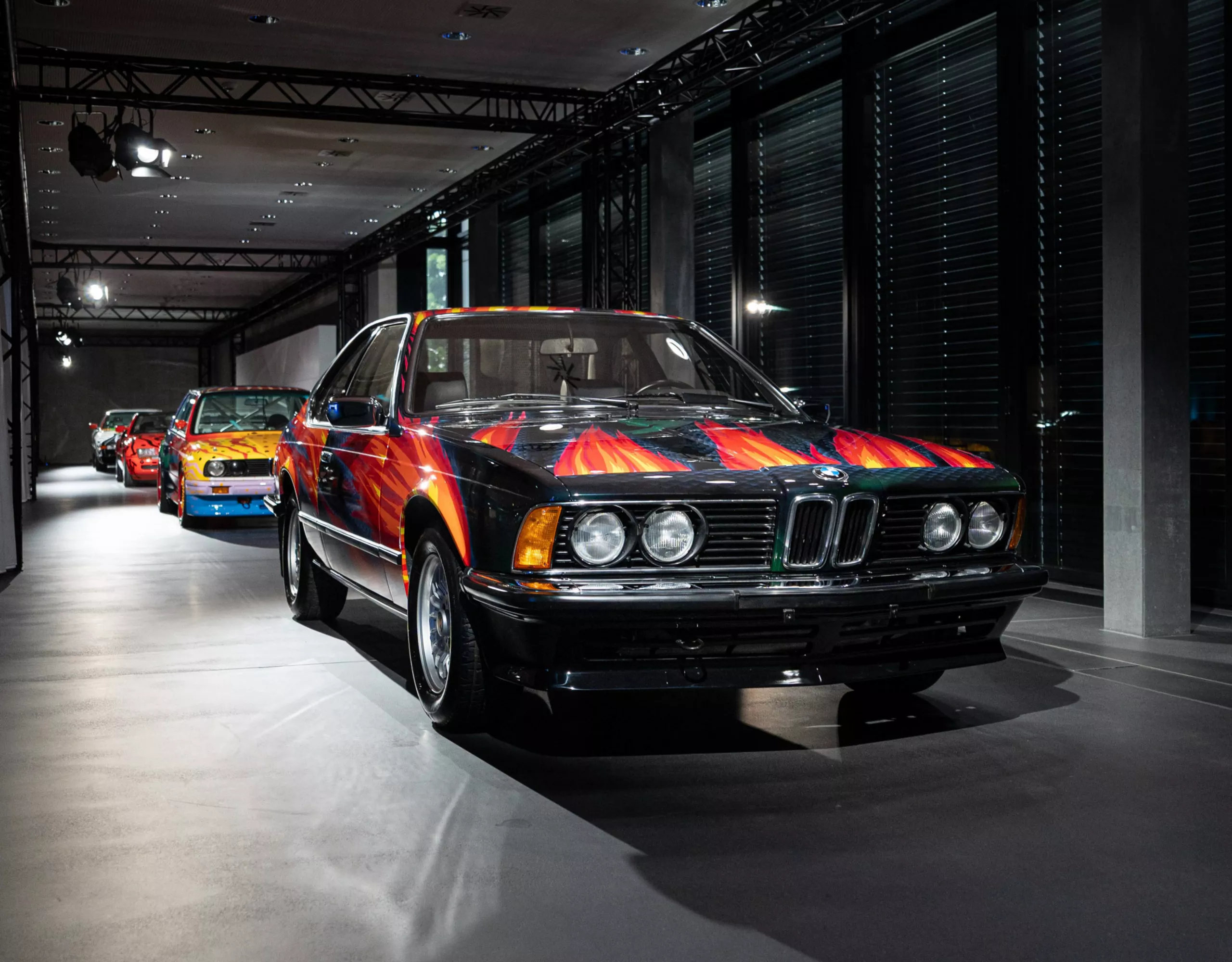 Vista frontale della BMW Art Car “Feuerfuchs auf Hasenjagd” di Ernst Fuchs con vernice nera e lingue di fuoco ardenti con altre Art Car sullo sfondo