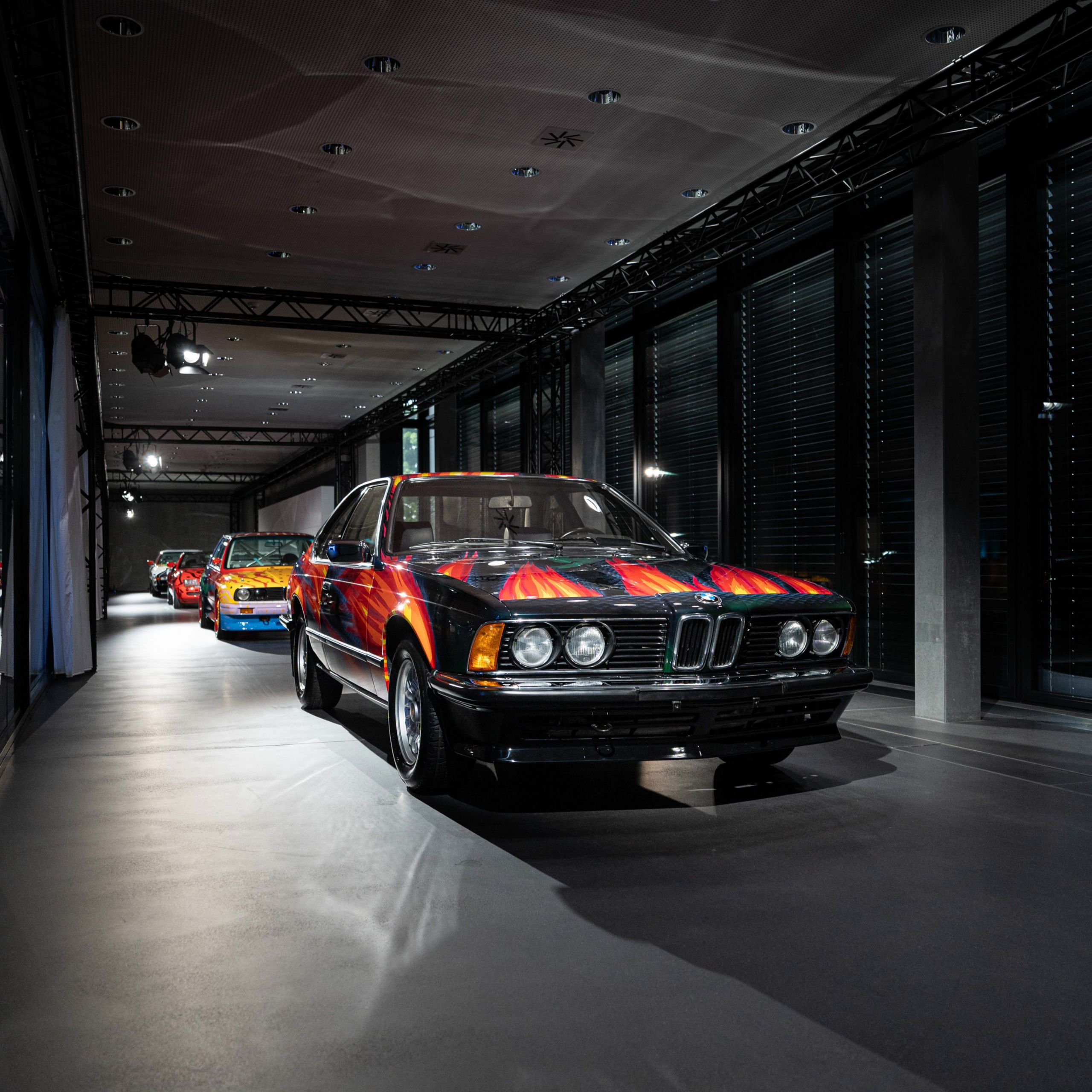 Vue de face de la BMW Art Car «Feuerfuchs auf Hasenjagd» d’Ernst Fuchs avec une peinture noire et des langues de feu flamboyantes, avec d’autres Art Cars en arrière-plan
