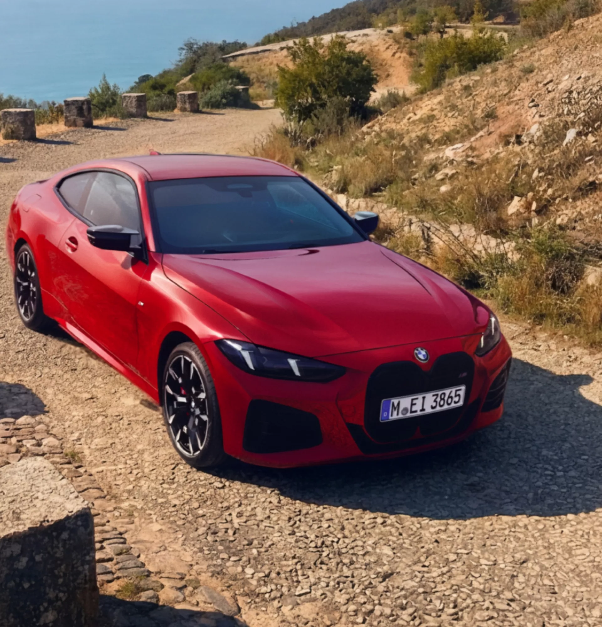 Ein BMW M440i xDrive Coupé in Rot steht auf einer schmalen Straße in einer trockenen, hügeligen Landschaft mit Felsen und wenig Vegetation. Das sportliche Fahrzeug wird aus leichter Vogelperspektive gezeigt und hebt sich durch seine auffällige Farbe klar von der natürlichen Umgebung ab. Die Szene vermittelt Dynamik und Performance in einer ruhigen, ländlichen Kulisse.