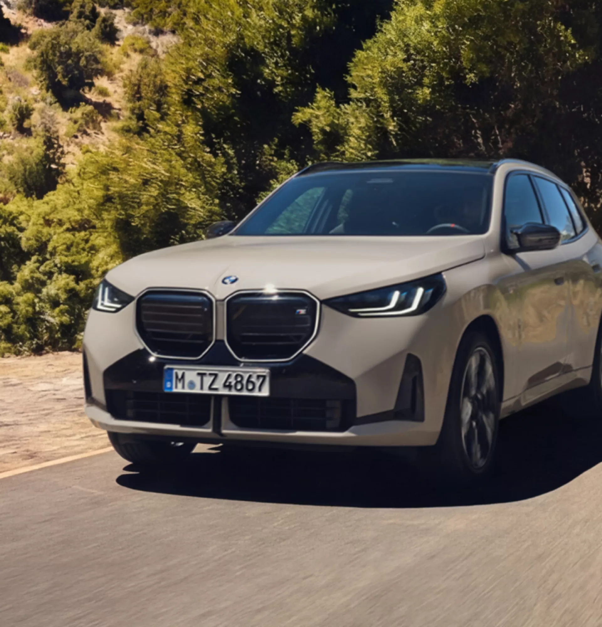 Ein BMW X3 M50 xDrive fährt auf einer kurvenreichen Straße durch eine hügelige Landschaft mit trockener Vegetation und Bäumen im Hintergrund. Das helle Fahrzeug hebt sich dynamisch von der natürlichen Umgebung ab und steht für Vielseitigkeit und sportliche Fahrleistung. Die Szene vermittelt Bewegung und Fahrspaß in einer sonnigen Outdoor-Kulisse.