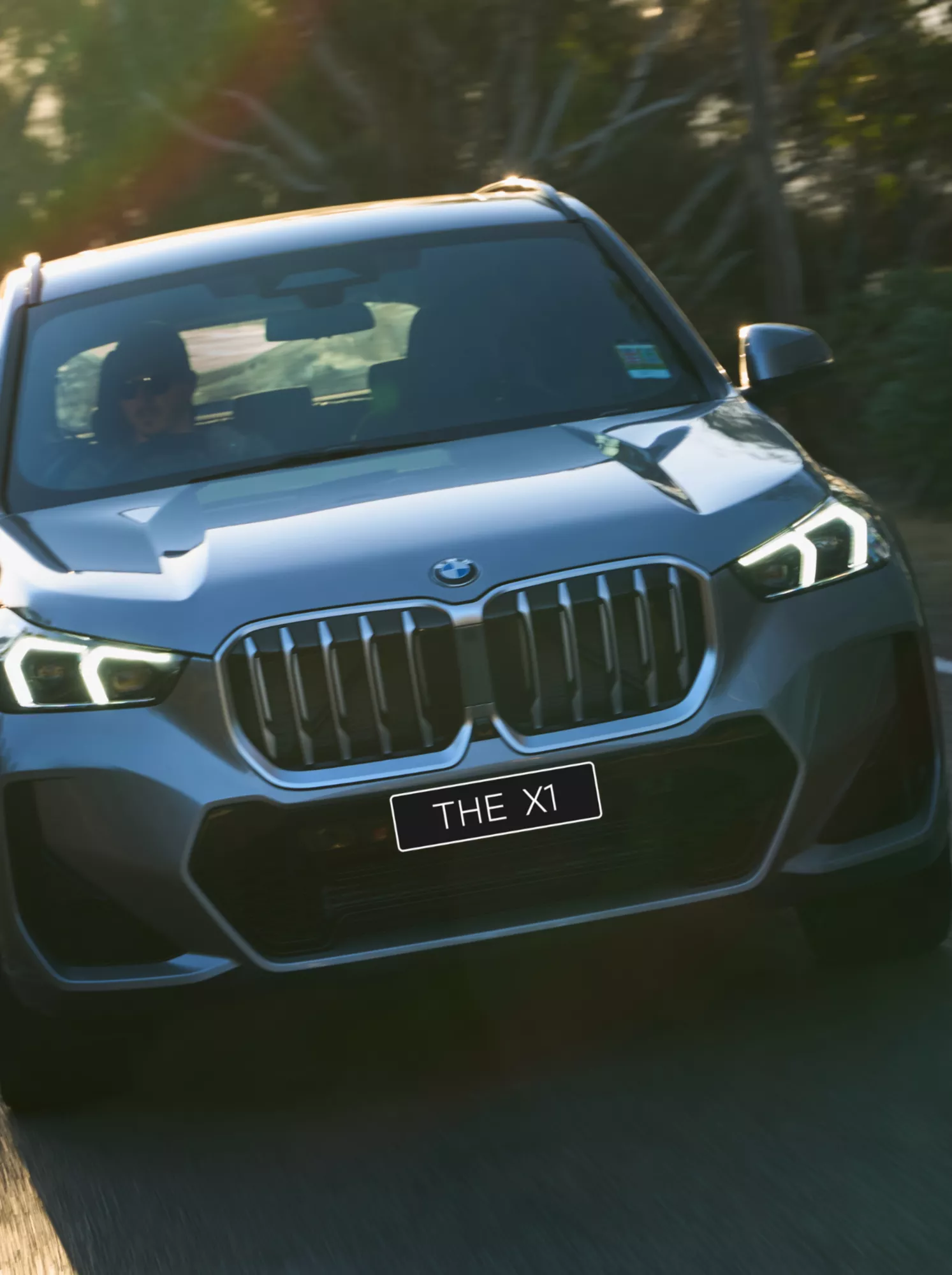  Front on view of a grey BMW X1 Plug-In Hybrid driving on the road with trees in the background