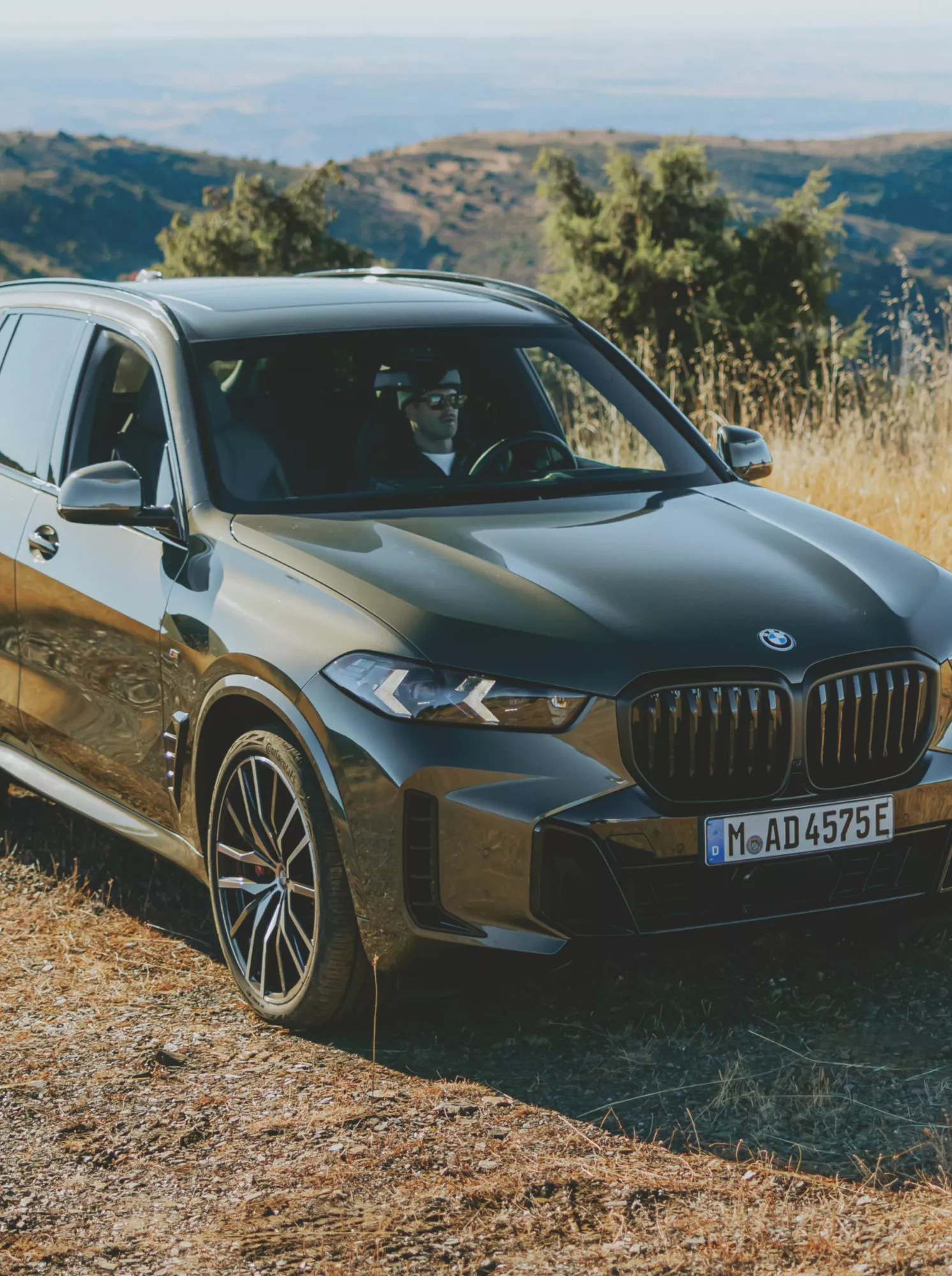 A dark grey BMW X5 plug-in hybrid SUV parked on a rugged dirt hillside surrounded by golden dry grass, with a sweeping panoramic valley and rolling hills in the background