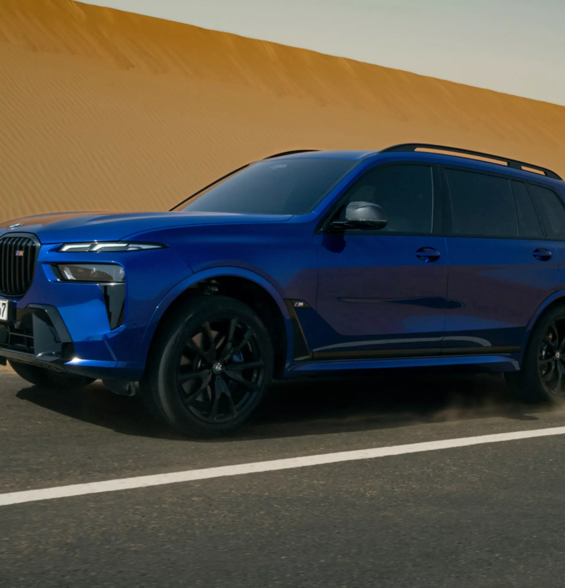 Ein blauer BMW X7 fährt auf einer Straße entlang einer großen Sanddüne, während etwas Staub von den Reifen aufgewirbelt wird.