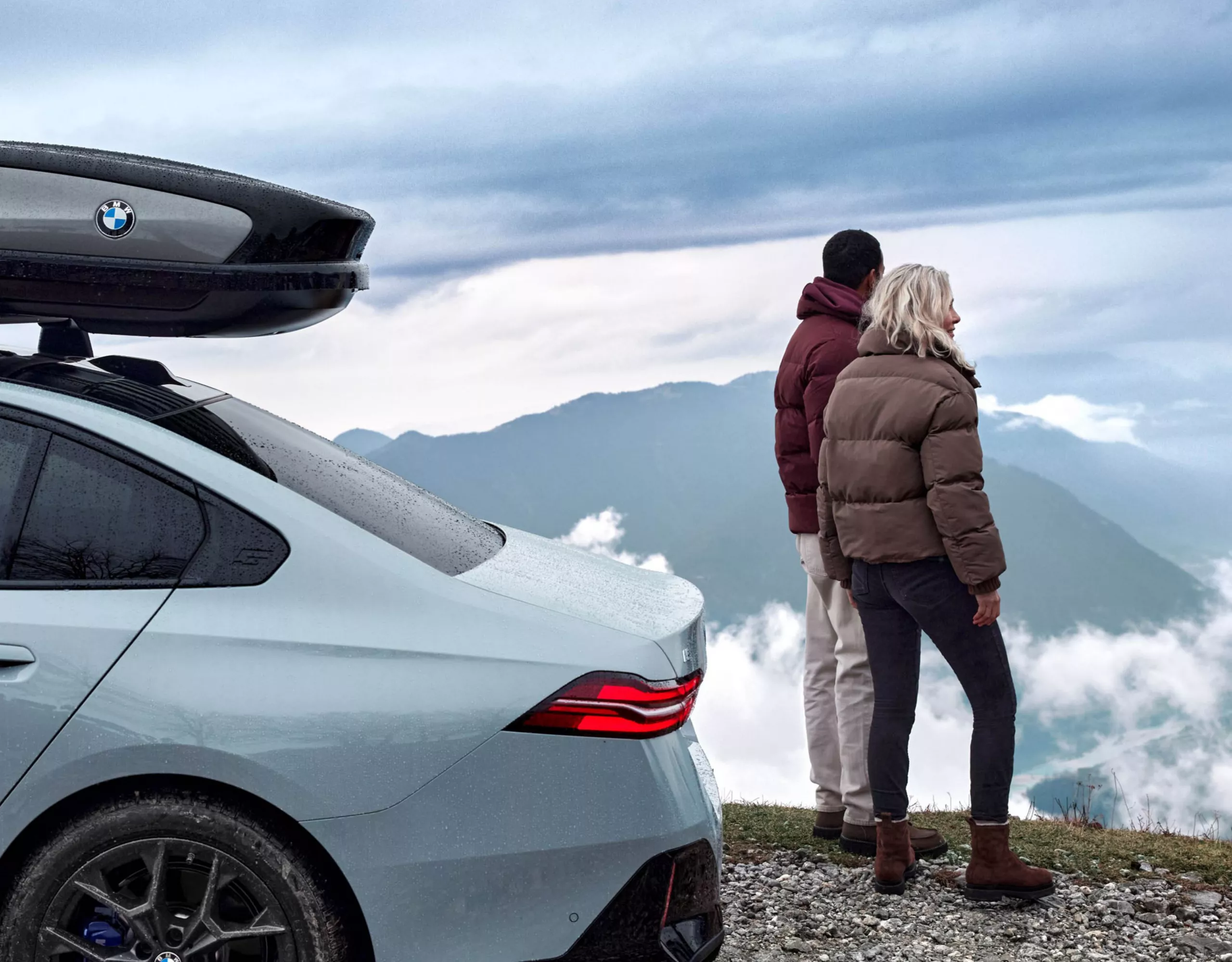 En BMW i5 Sedan med takbox parkerat högt uppe på ett berg. Bergsmassiv och människor i bakgrunden.