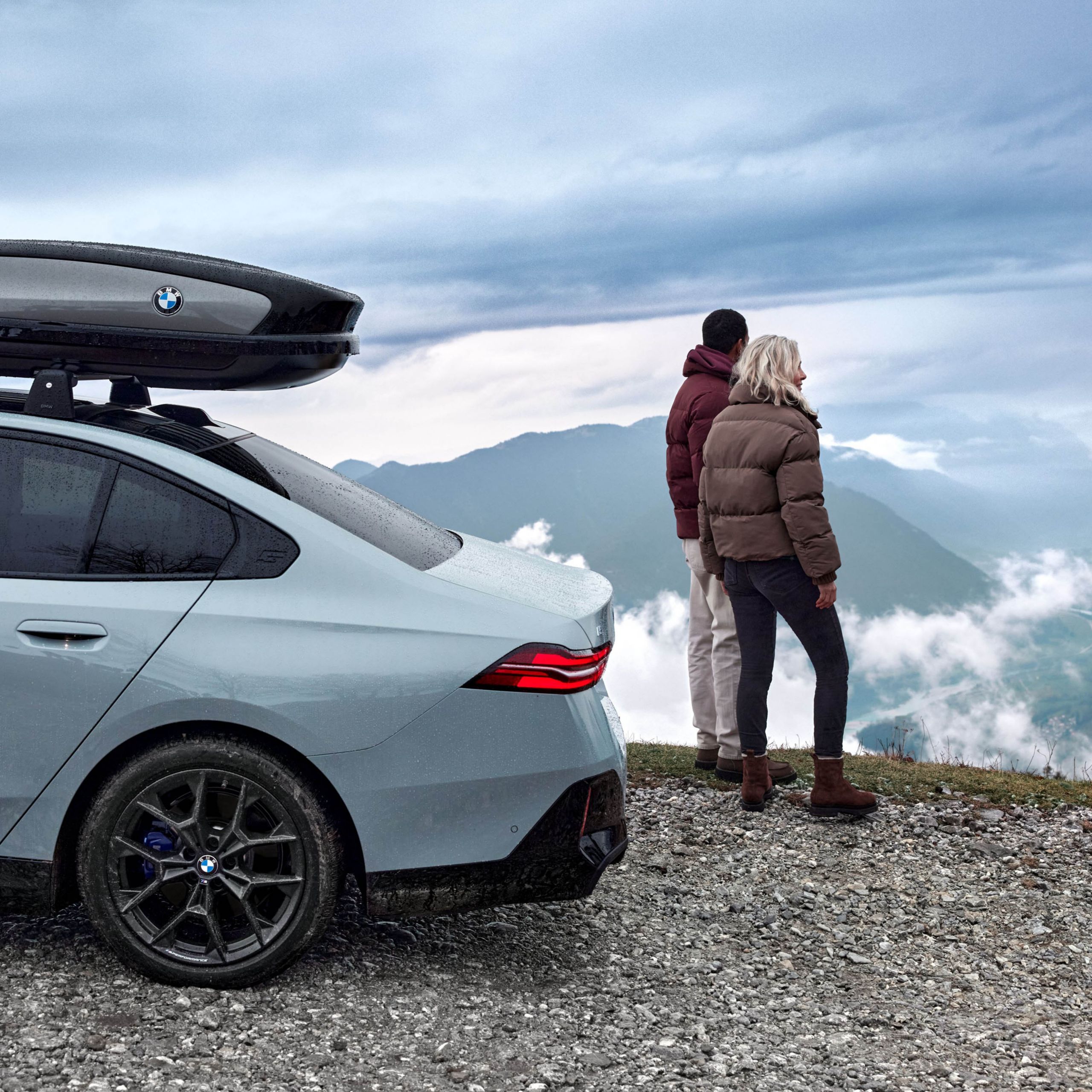 En BMW i5 Sedan med takbox parkerat högt uppe på ett berg. Bergsmassiv och människor i bakgrunden.