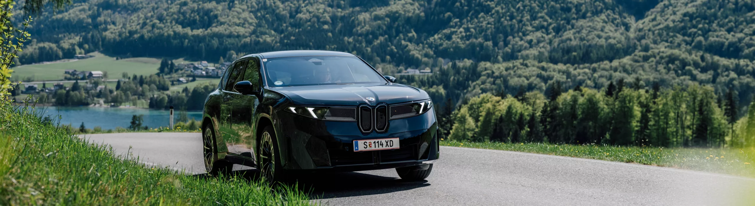 Ein dunkelblauer BMW iX3 (NA5) fährt über eine sonnige Landstraße in einer grünen Landschaft.