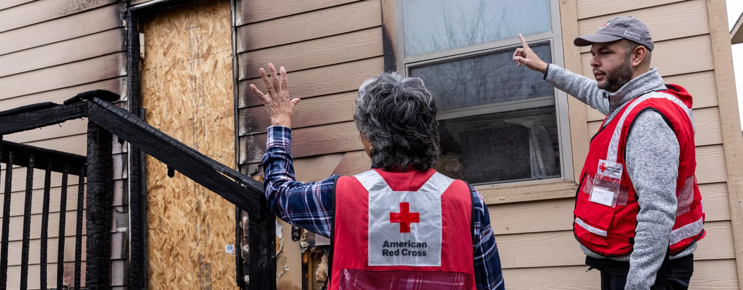 Two Red Cross workers gesturing and looking at a house with fire damage