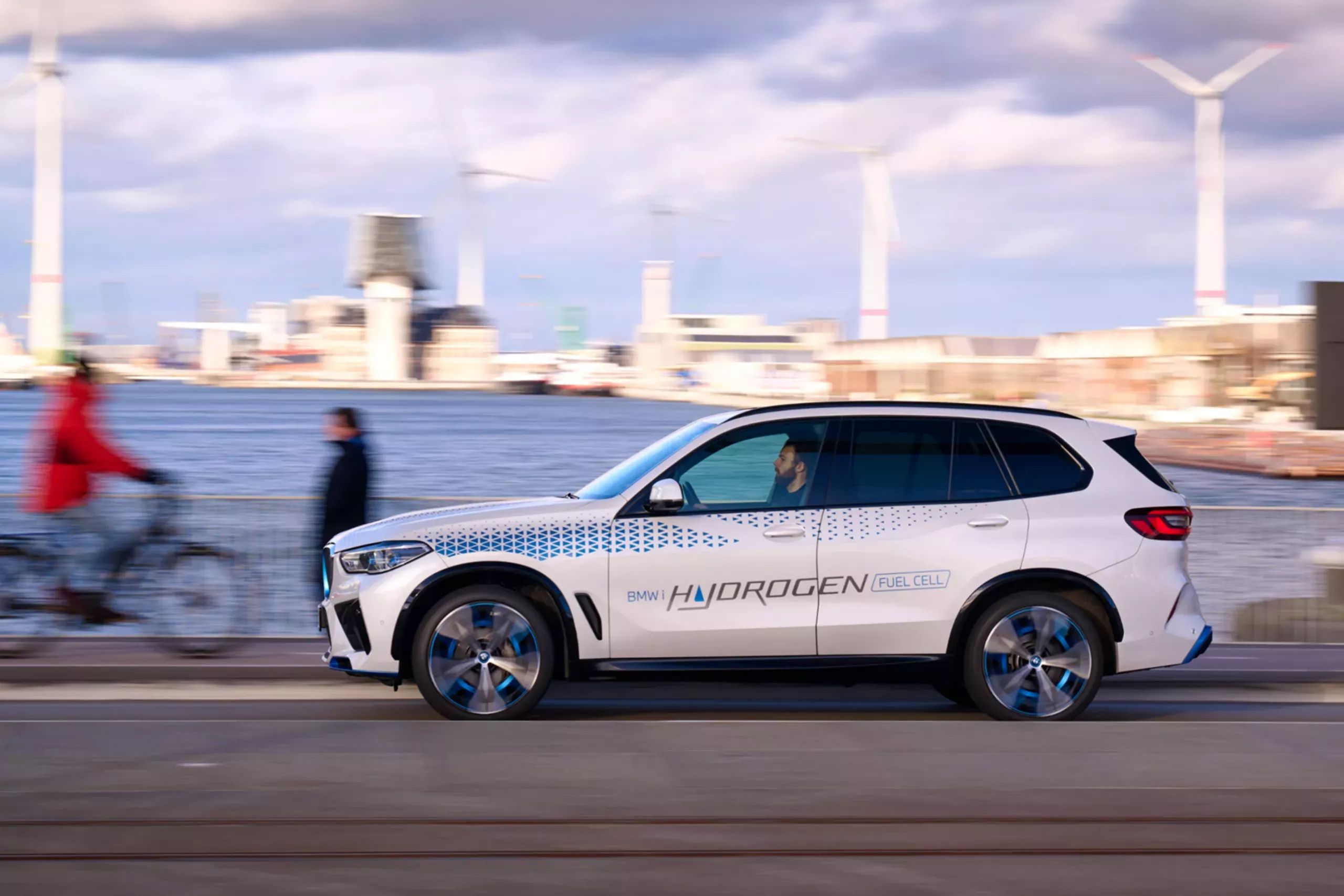 The BMW iX5 Hydrogen concept vehicle drives on a road adjacent to a body of water with wind turbines in the background