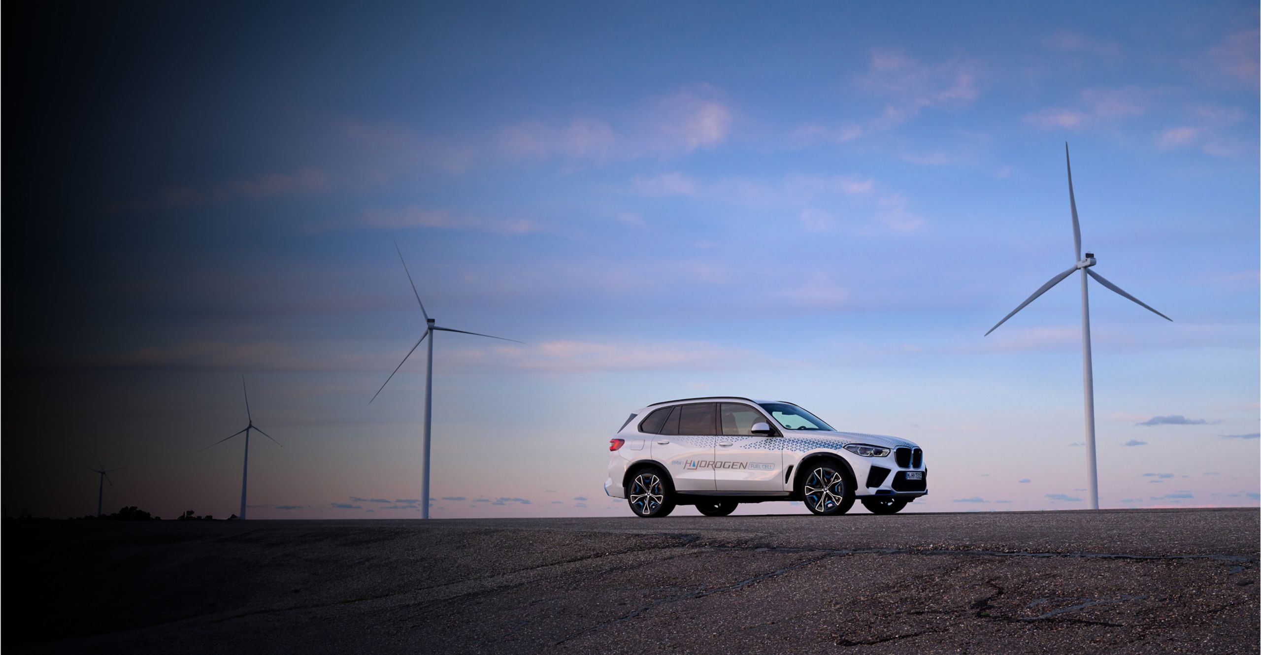 The BMW iX5 Hydrogen concept vehicle sits parked on a road with two modern windmills in the background