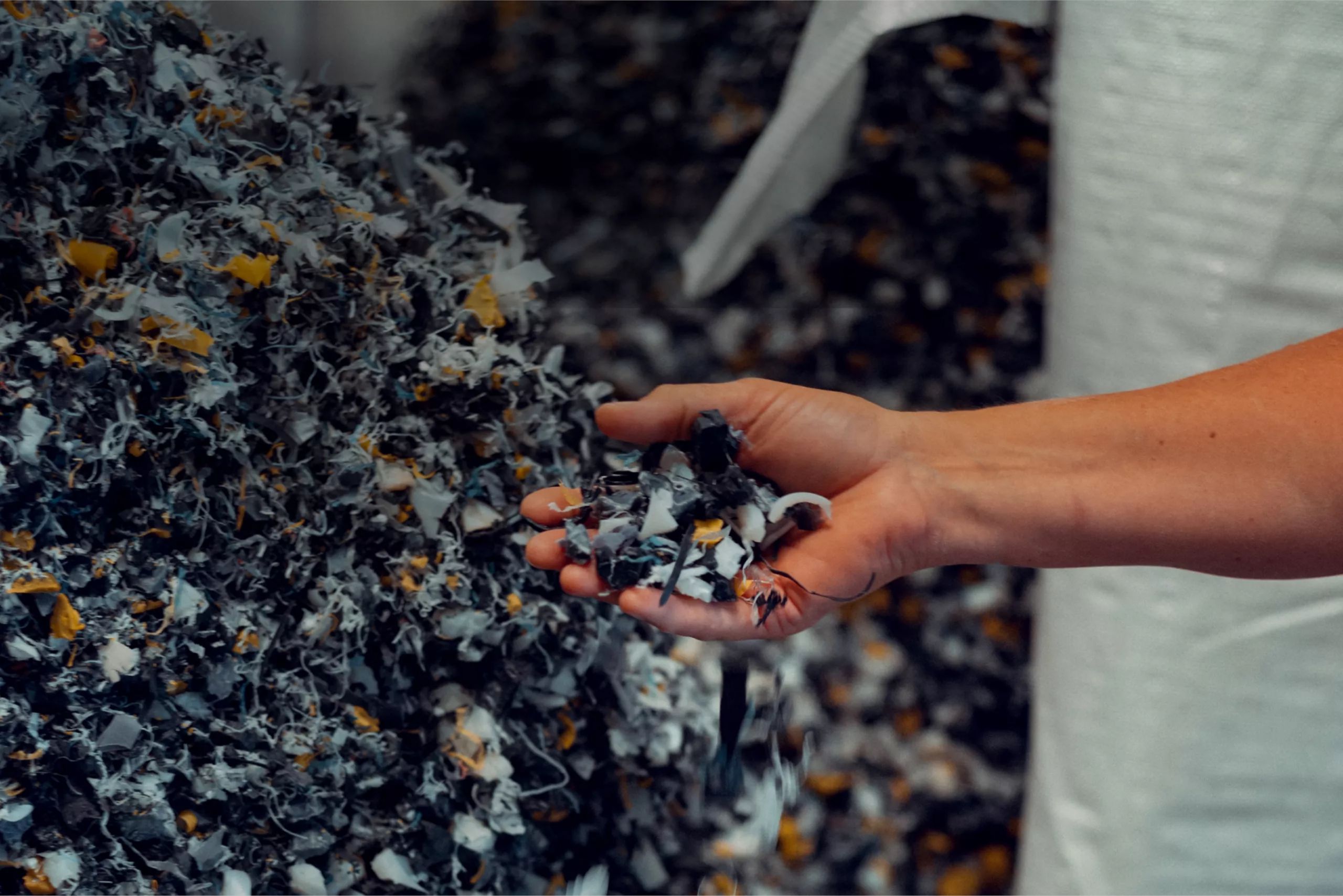 A person's hand picking up a handful of recycled material, with a large pile of recycled material in the background