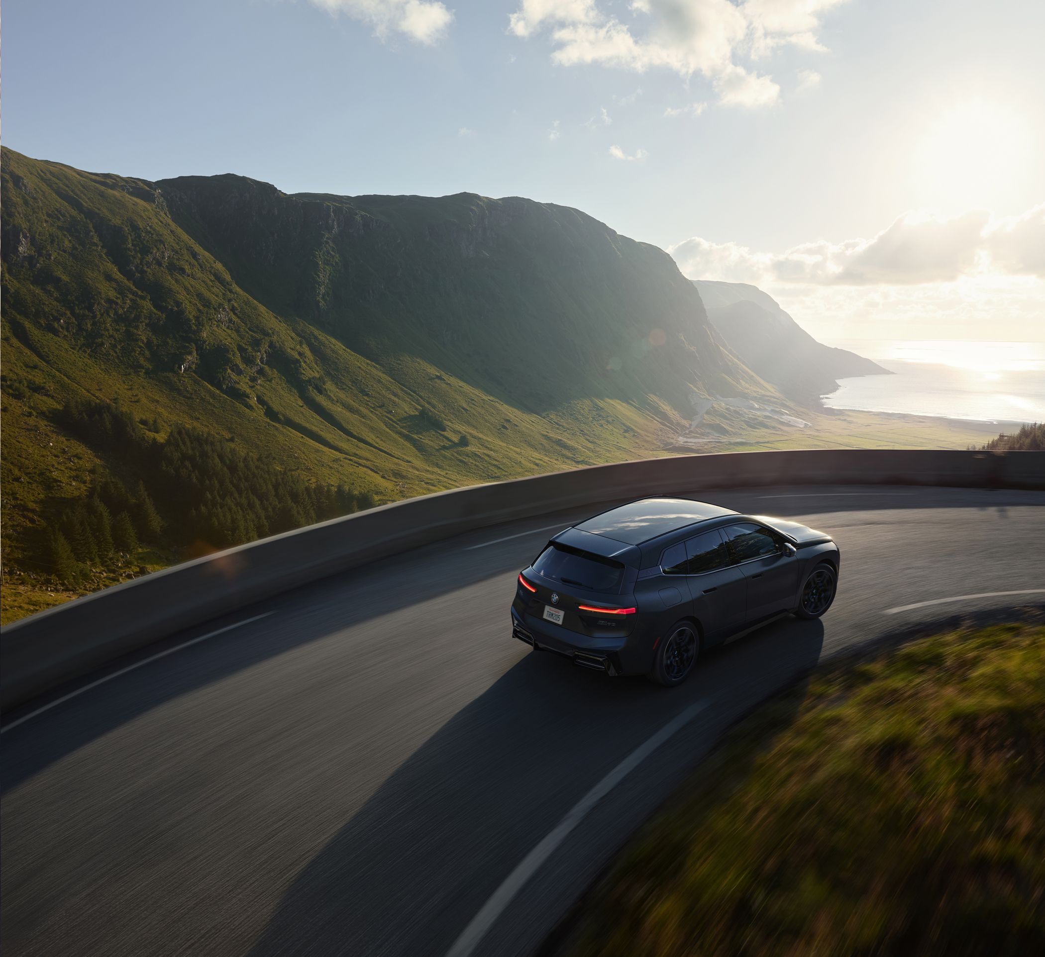 A BMW iX drives on a curving road with a scenic coastal landscape in the background