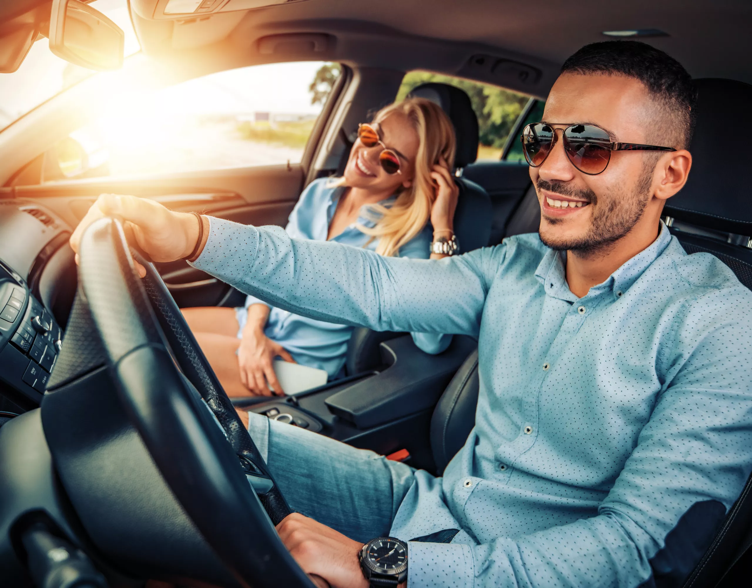 A couple smiling as they're driving with the sun in the background shining through the window