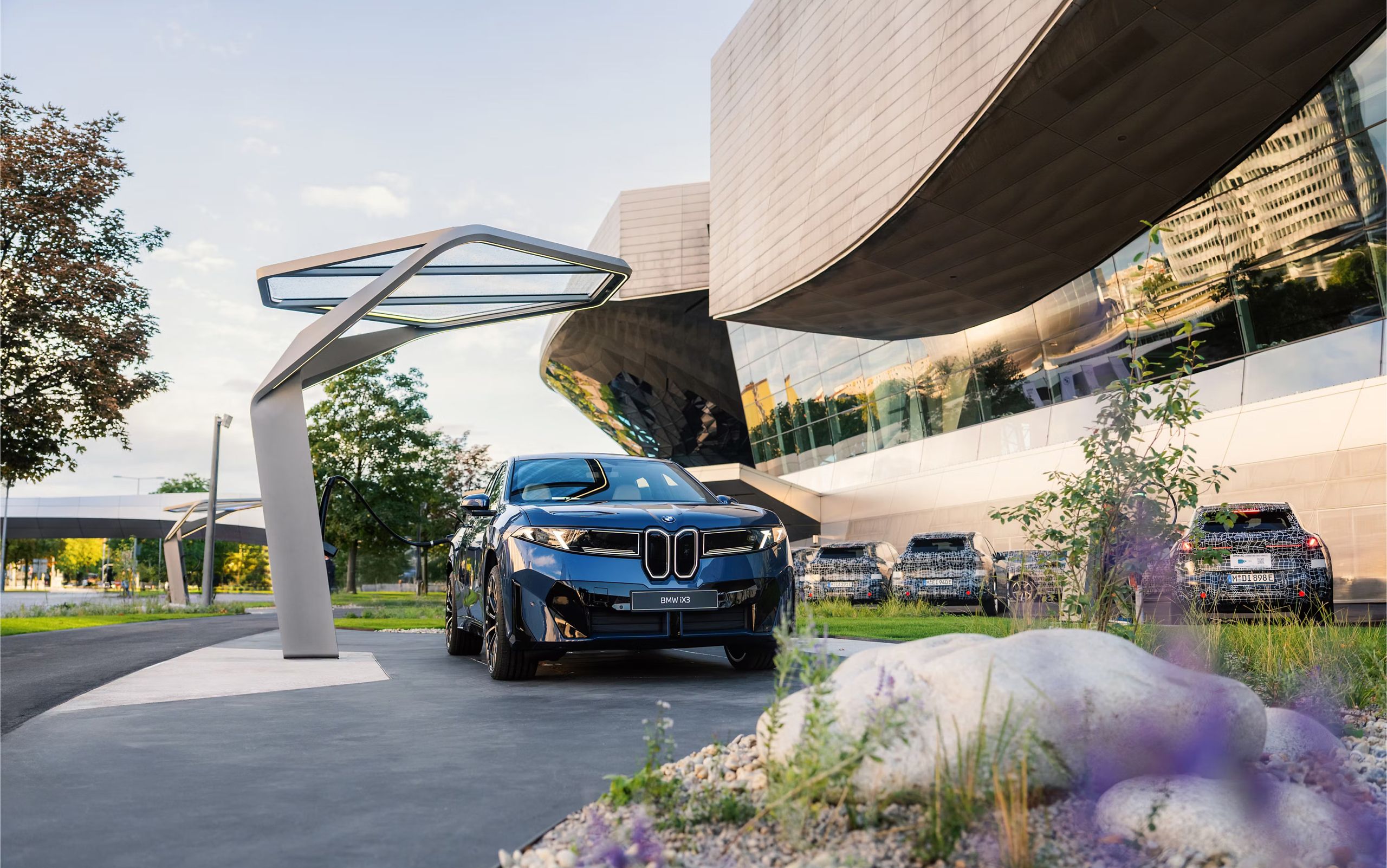 A BMW iX3 sits parked outside a modern looking building with beautiful landscaping