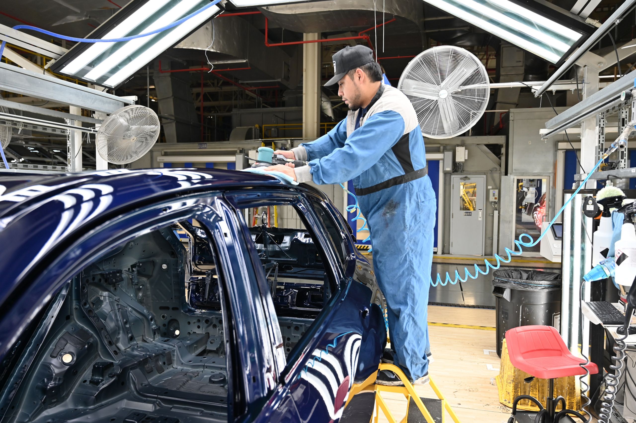 A man stands on a ladder, polishing the body of a vehicle in a factory