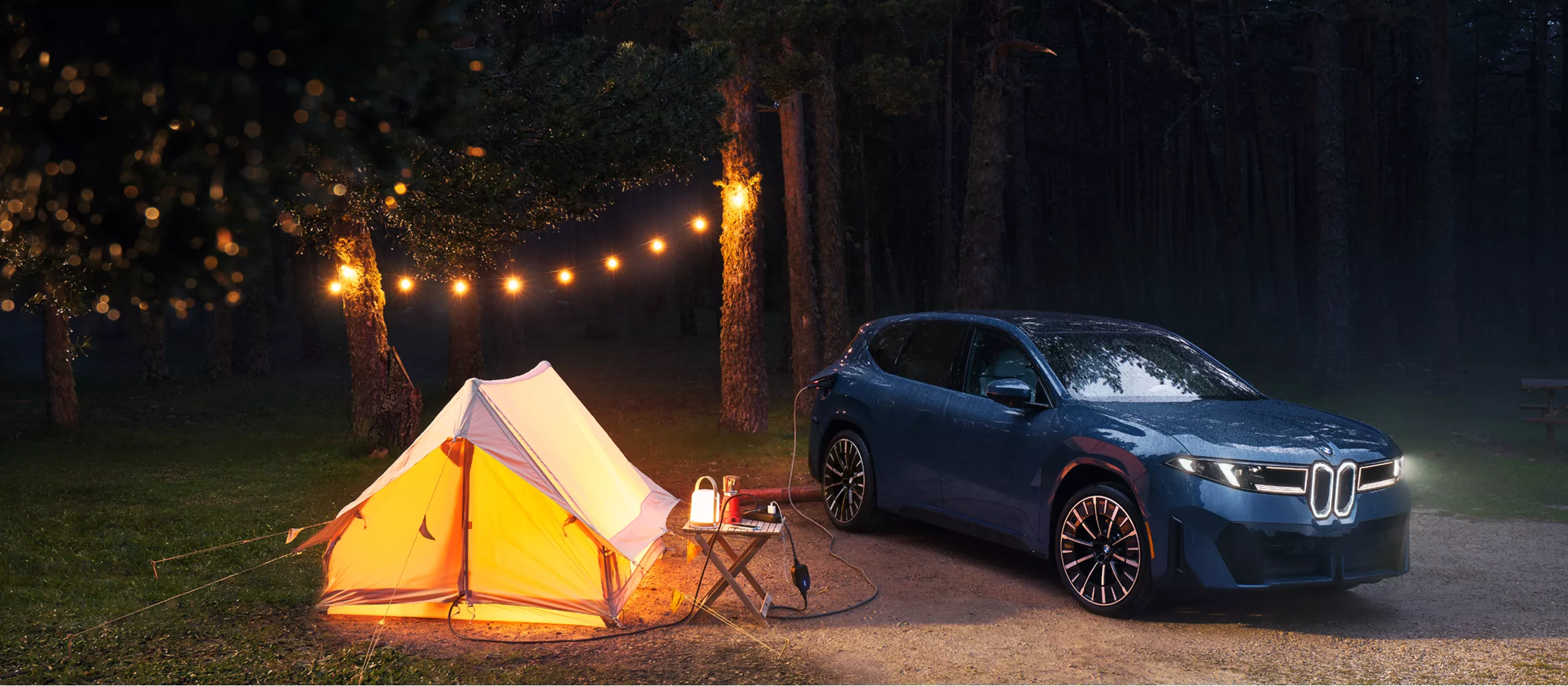 A BMW iX3 is parked at a campground while its charging cable leading into the tent to provide power 