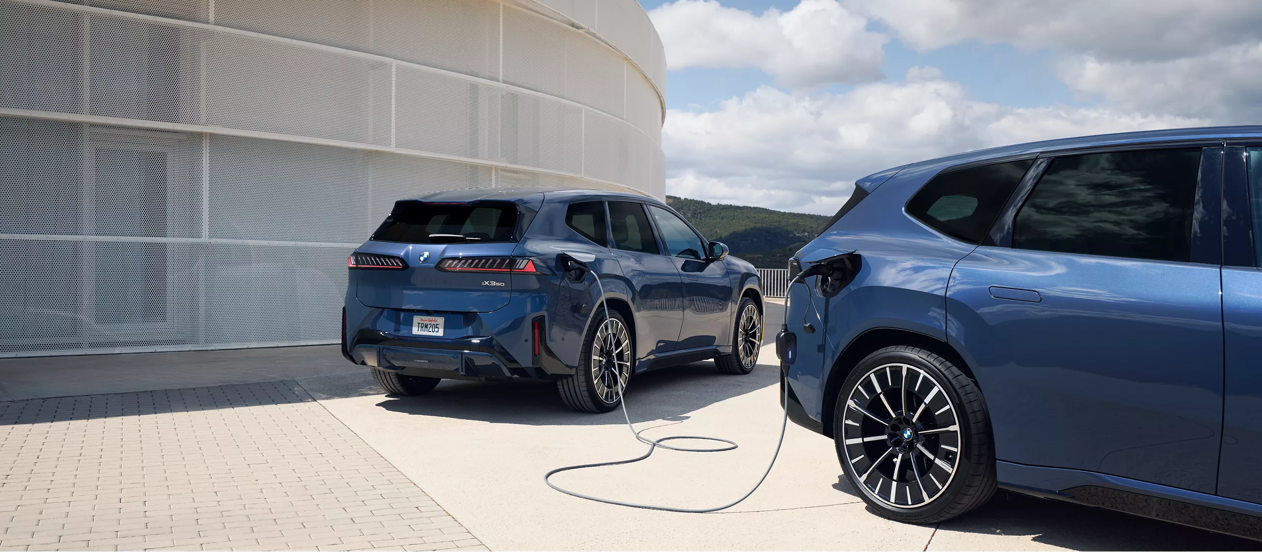 Two BMW iX3s are parked next to each other, one providing power to the other via its charging cable. 