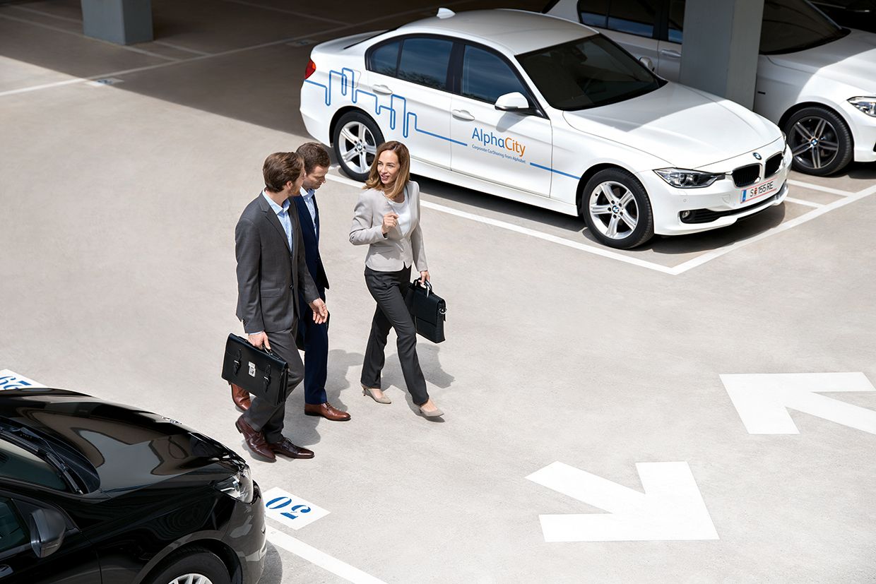 Drei Personen gehen auf einem asphaltierten Parkplatz an einem weißen BMW mit Alphabet-Beklebung vorbei.