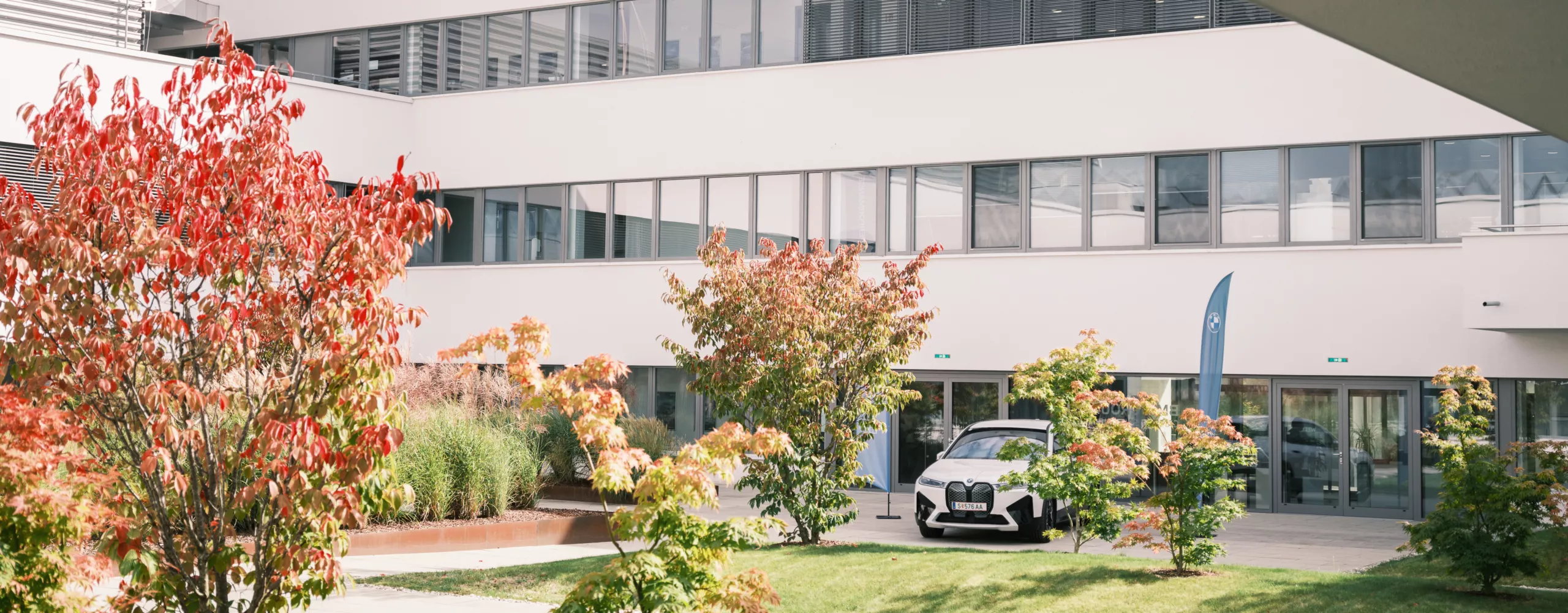 Im Innenhof des BMW Group Standorts Campus Nord steht ein weißer BMW iX zusammen mit zwei blauen BMW-Beachflags. Der Innenhof ist mit einigen Sträuchern begrünt.