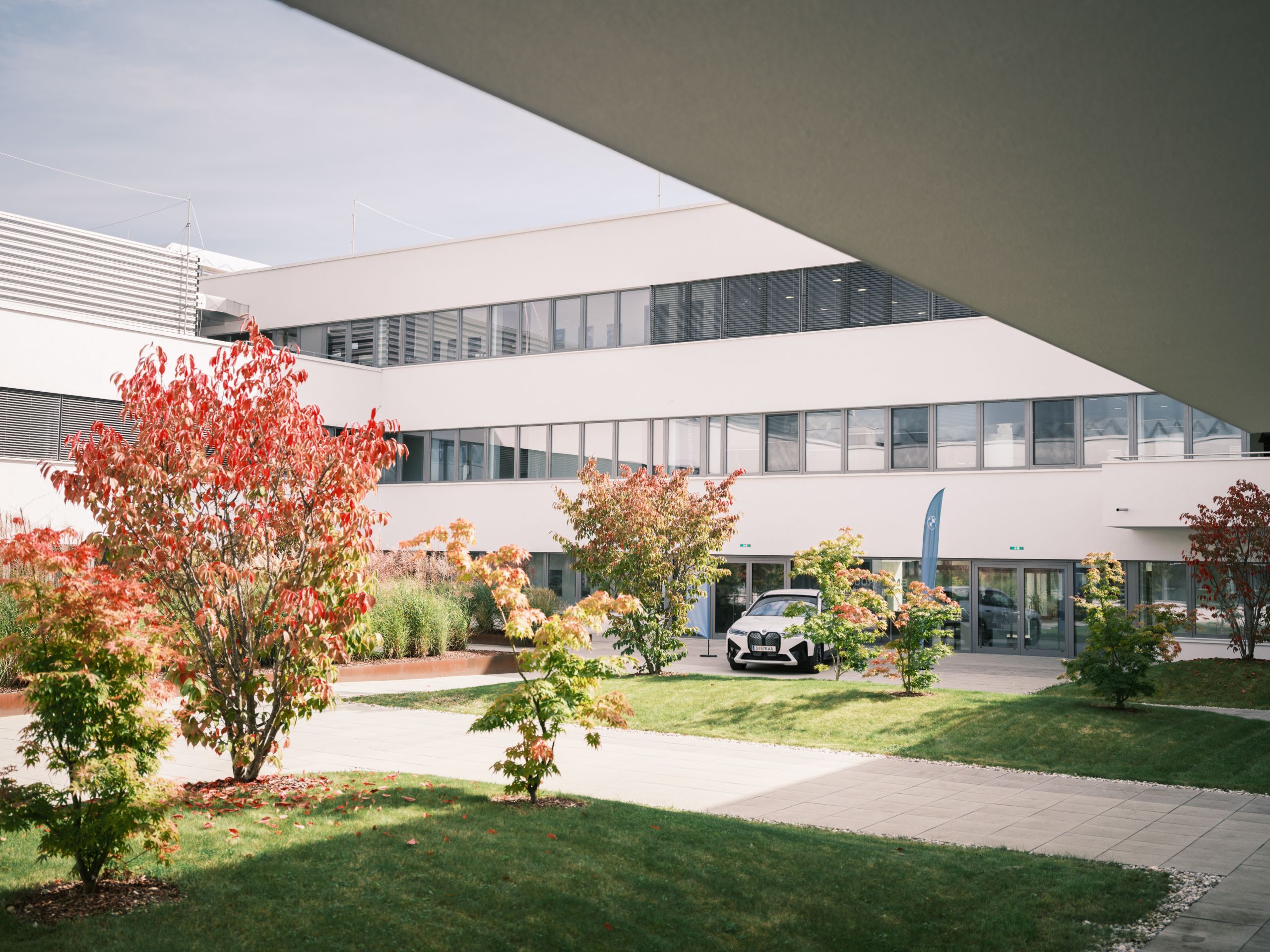 Im Innenhof des BMW Group Standorts Campus Nord steht ein weißer BMW iX zusammen mit zwei blauen BMW-Beachflags. Der Innenhof ist mit einigen Sträuchern begrünt.